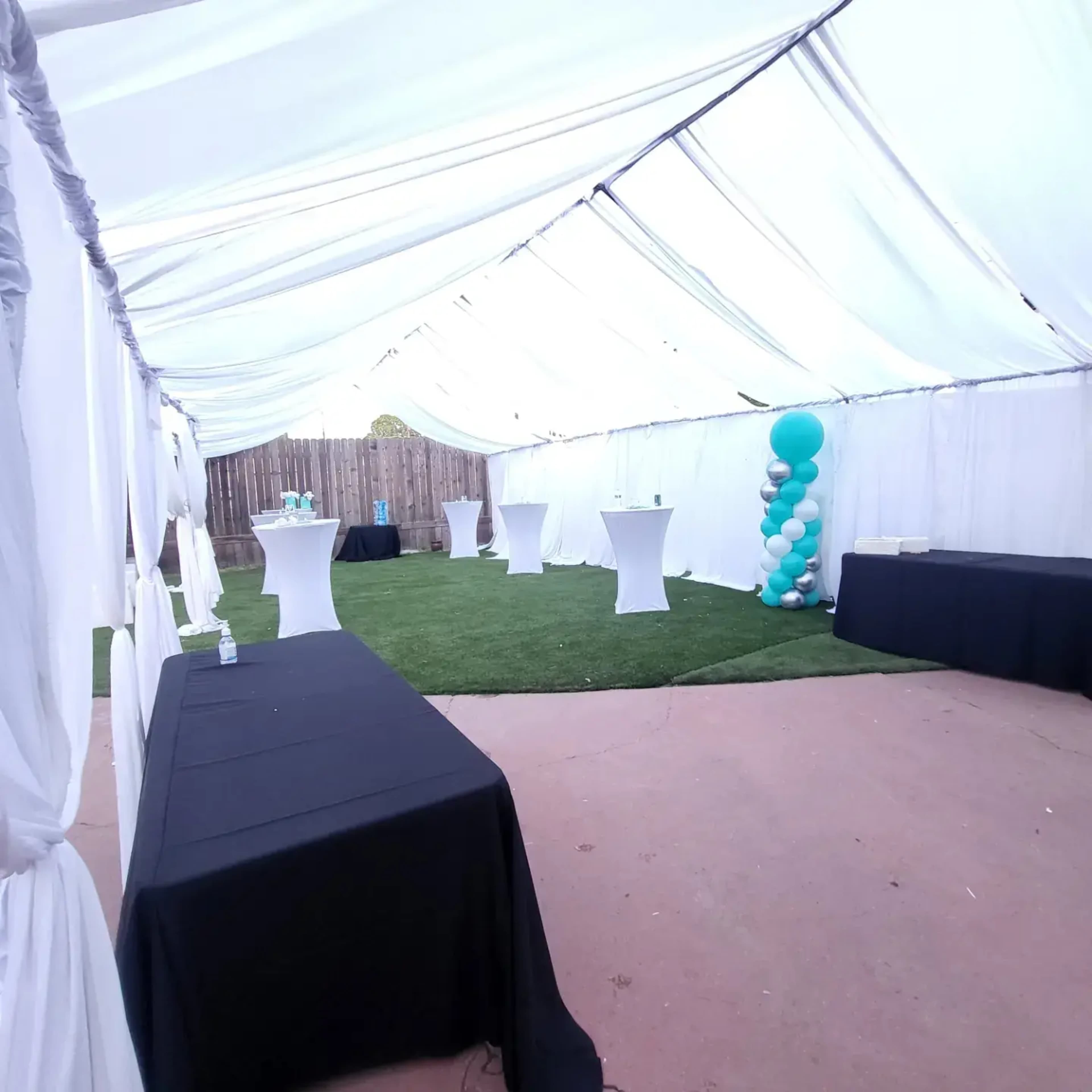 A large tent with white walls covers a grassy area, featuring several tables and a balloon decoration in teal and white.
