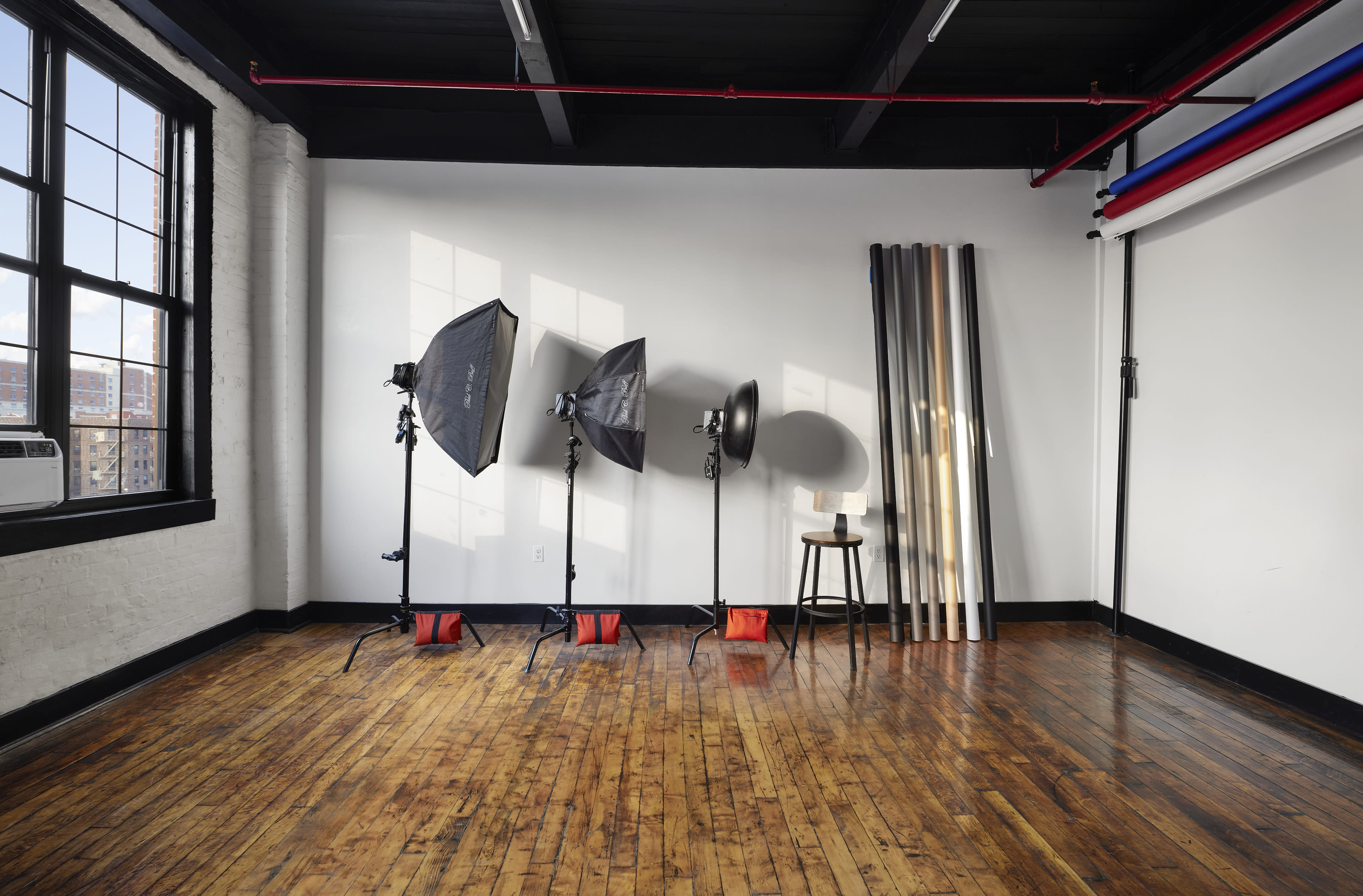 A photography studio featuring three light stands with softboxes, a single light source, a stool, and several rolls of backdrop material against a wall.