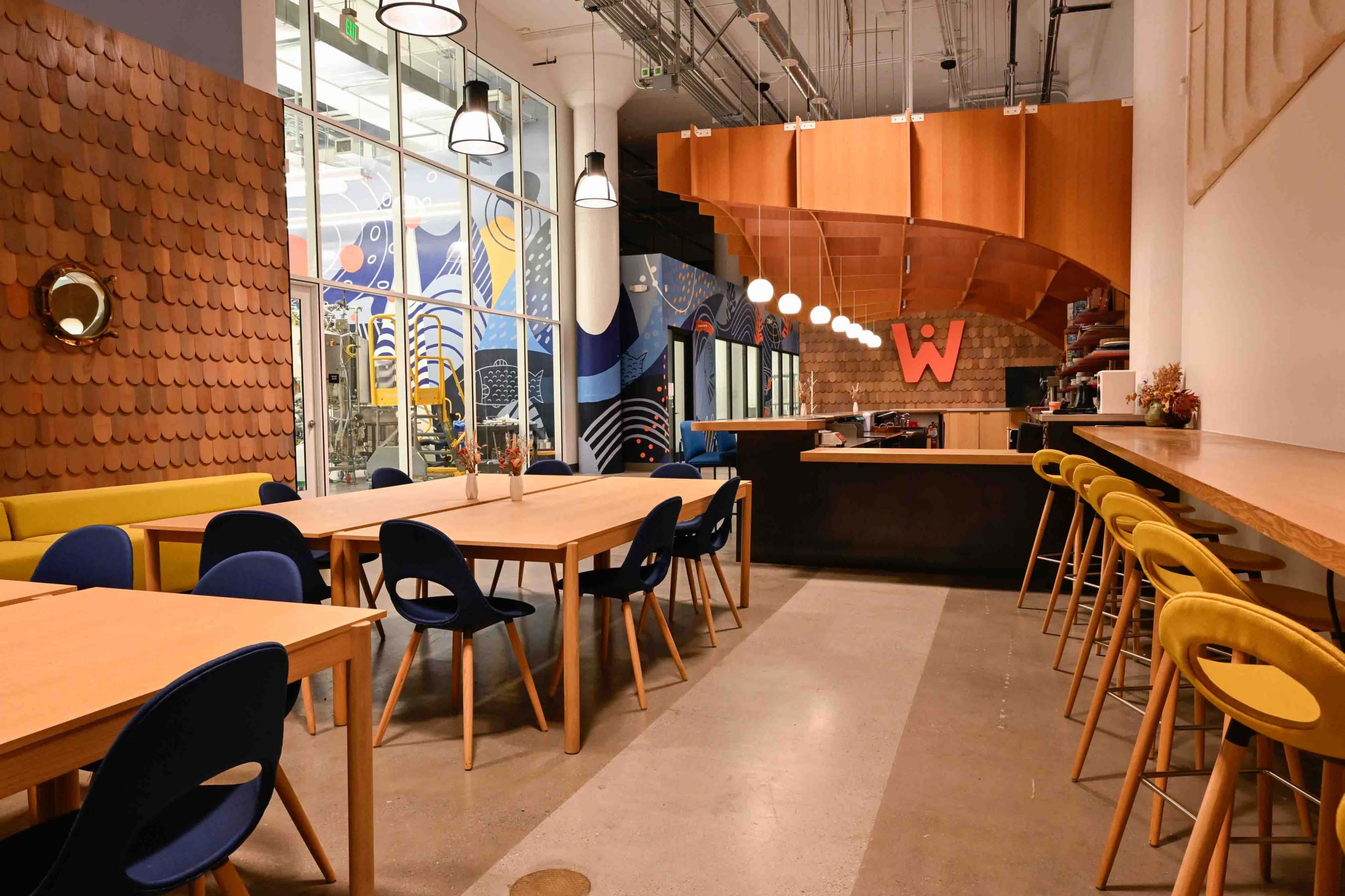 The image shows a modern, spacious dining area featuring wooden tables, colorful chairs, and a large mural on the wall.
