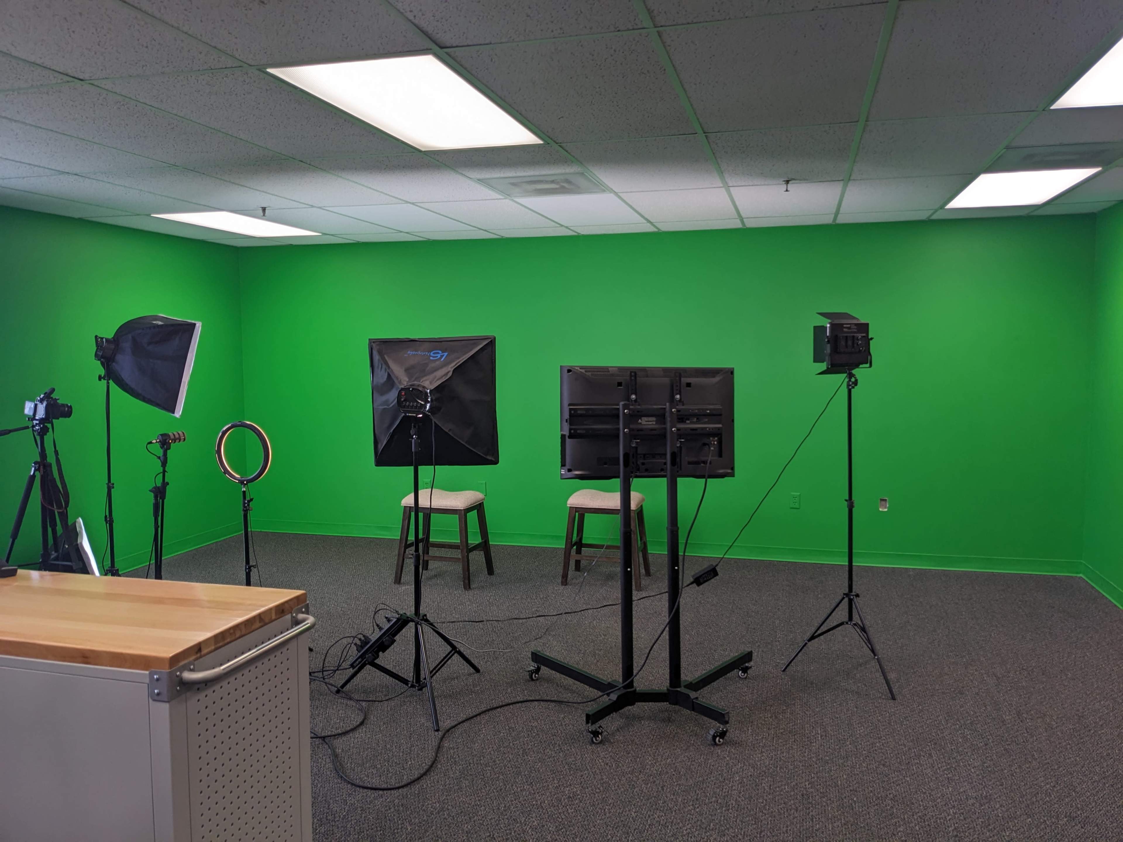 The image shows a recording studio with a bright green backdrop, two chairs, and several lights set up for video production.