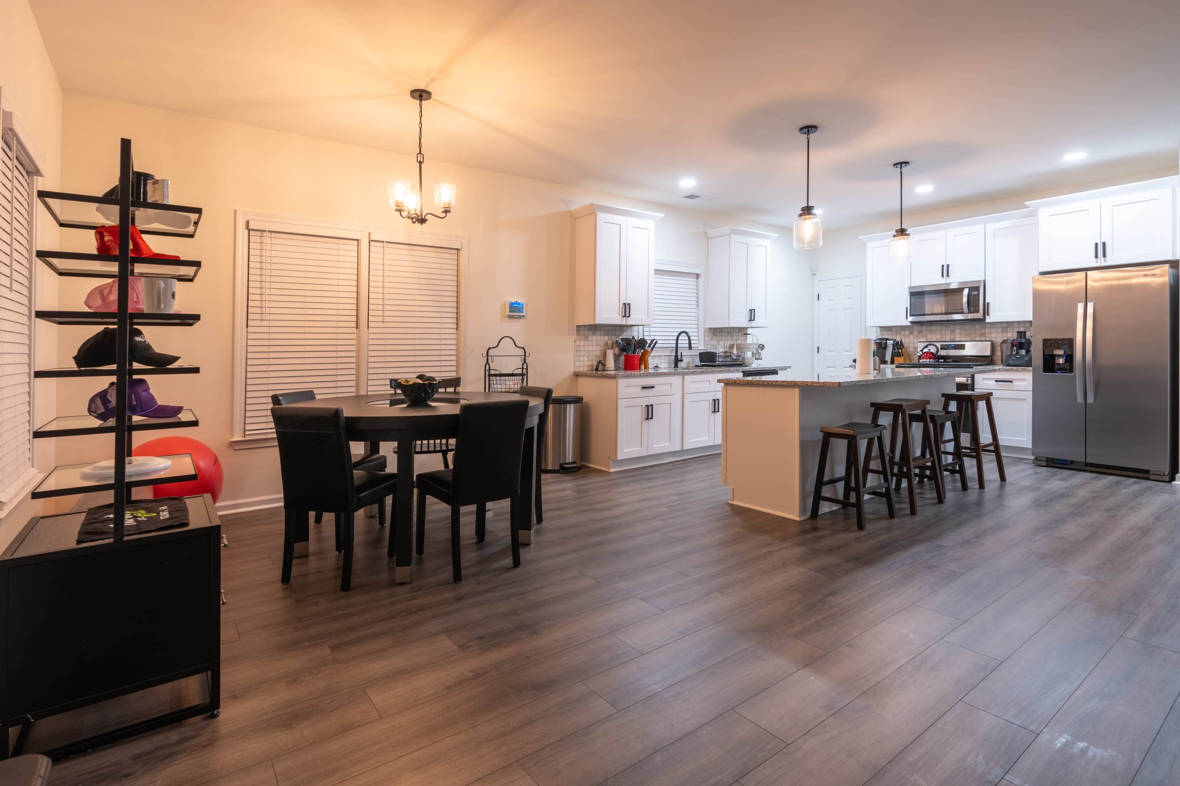 A modern kitchen and dining area features white cabinets, a dark island with bar stools, a dining table with four chairs, and various kitchen appliances.