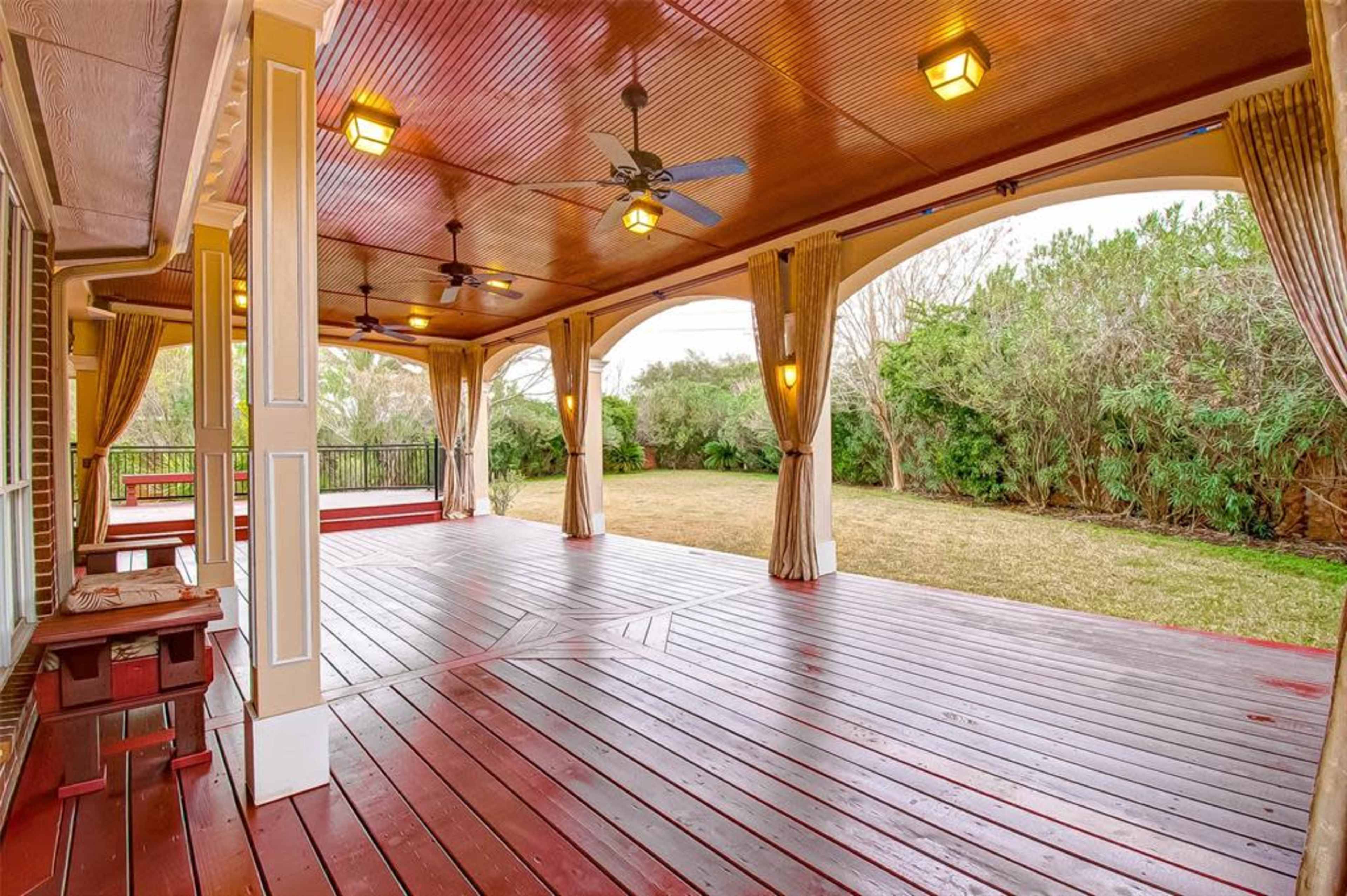 A large, covered patio with wooden flooring, ceiling fans, and curtains, overlooking a grassy backyard.