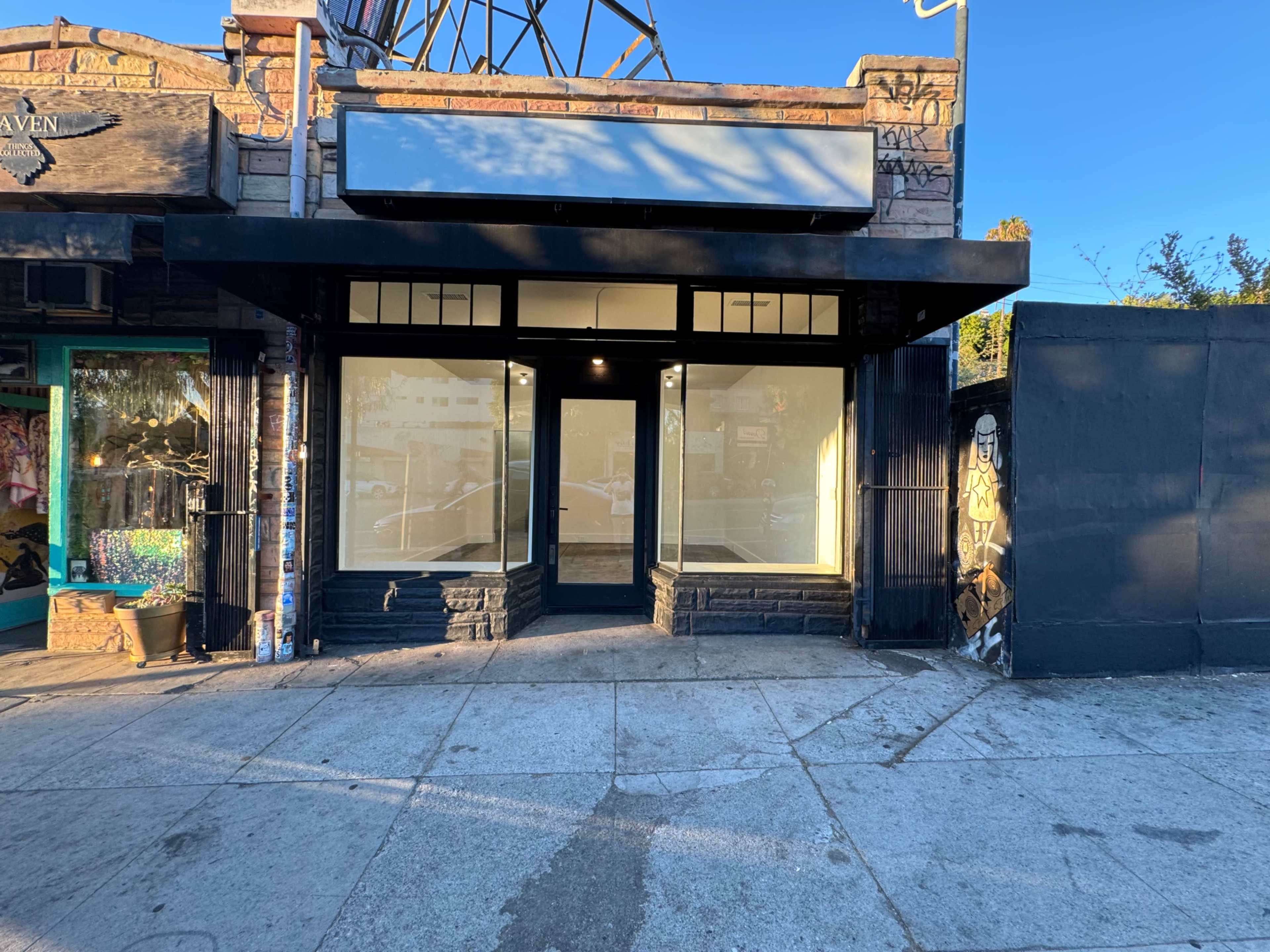 A storefront with large windows and a blank sign, located on a sidewalk adjacent to a graffiti-covered wall.