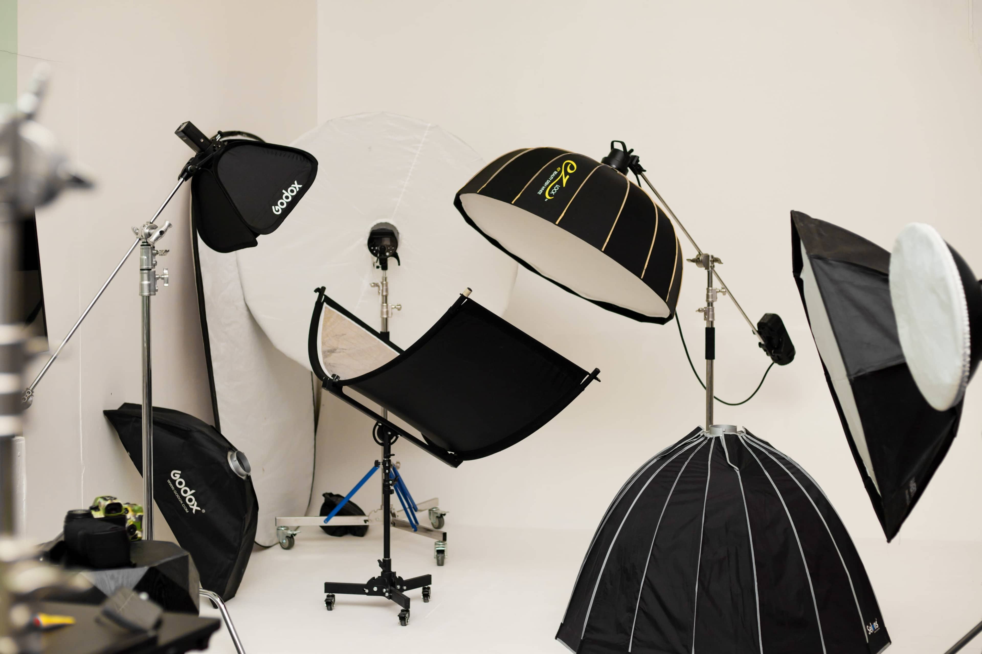 The image shows a photography studio setup featuring various softboxes and light reflectors arranged around a neutral background.