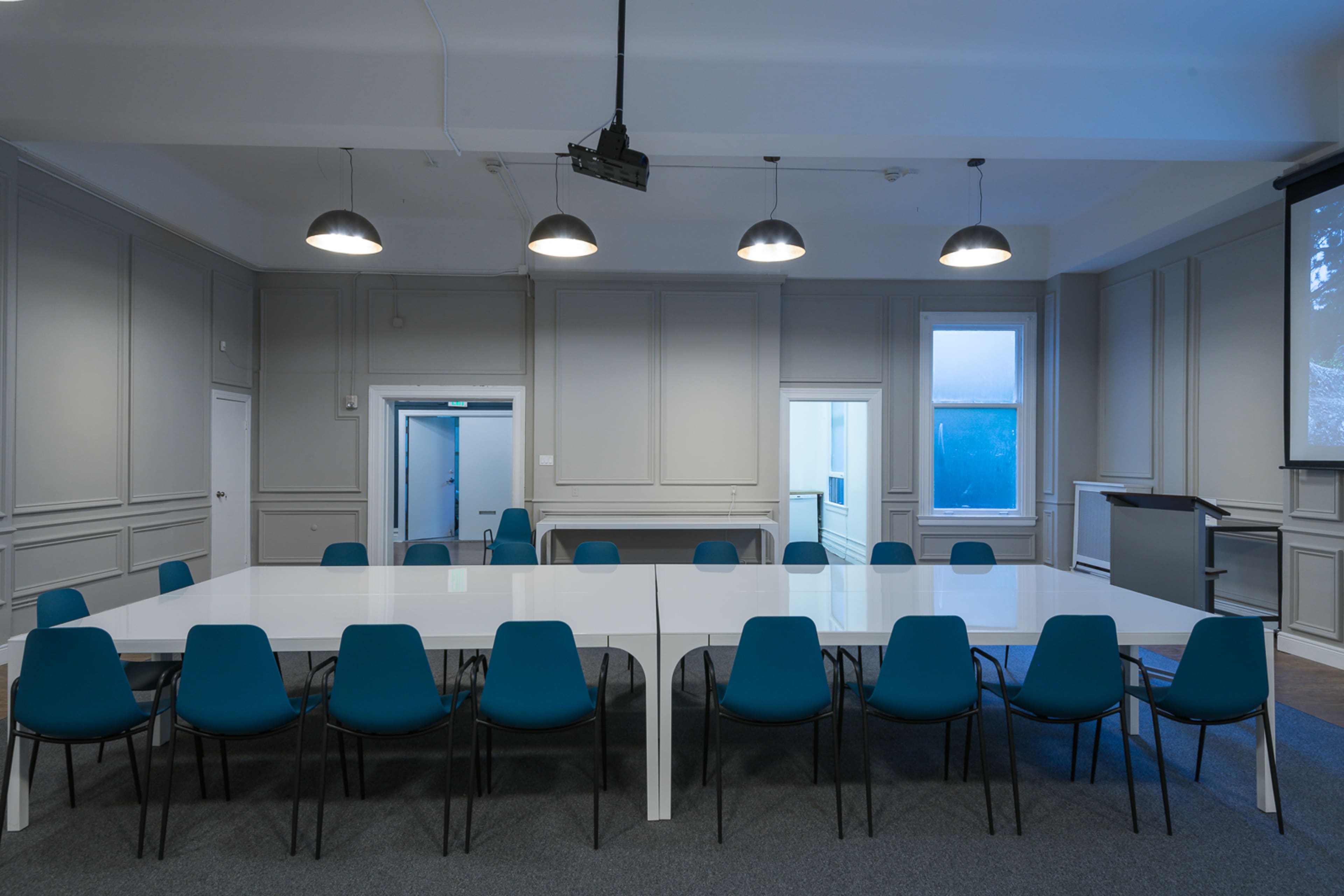 A spacious conference room features a large white table surrounded by blue chairs, with a projector and windows displaying natural light.