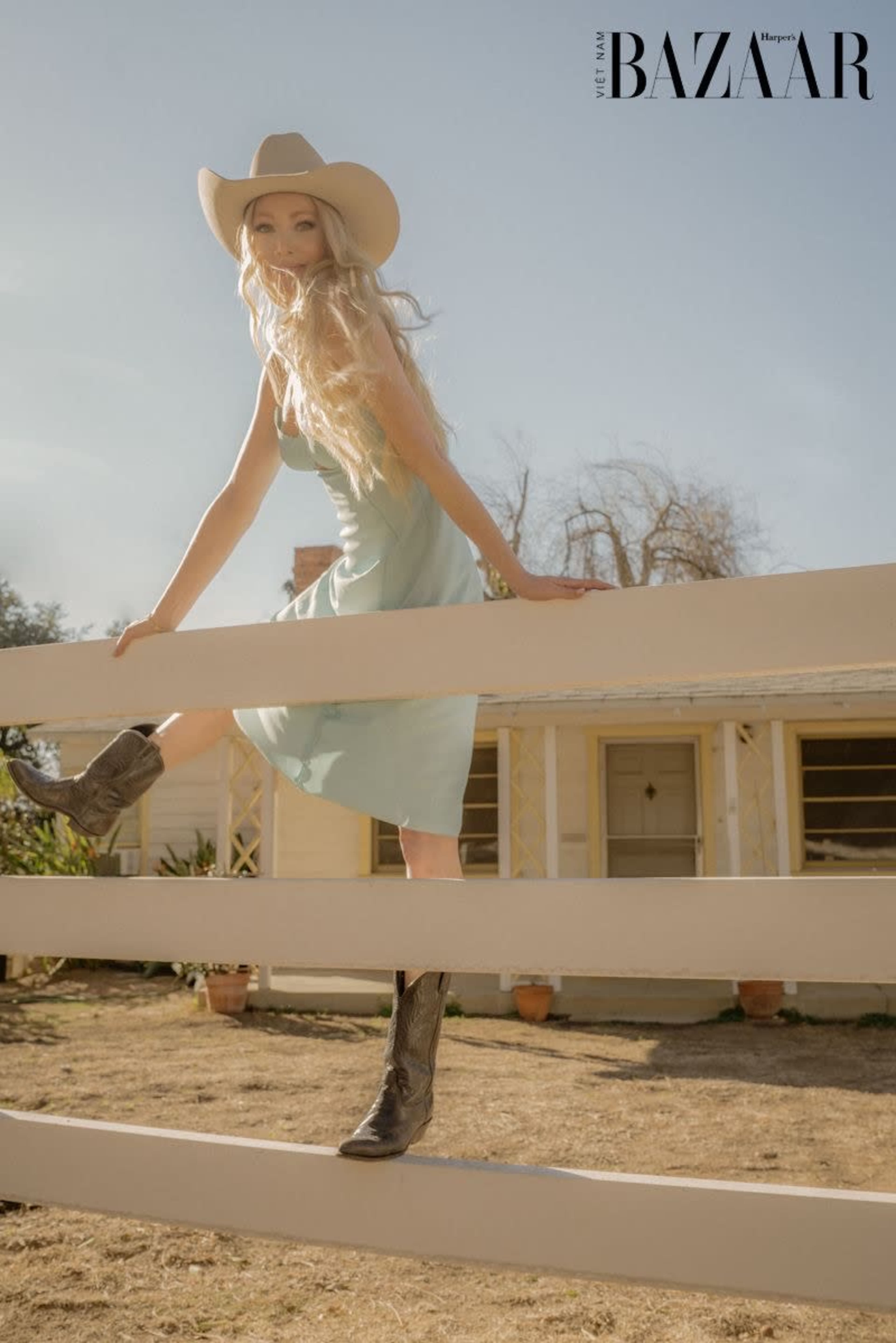 A woman in a light blue dress and cowboy boots is perched on a wooden fence in front of a house.
