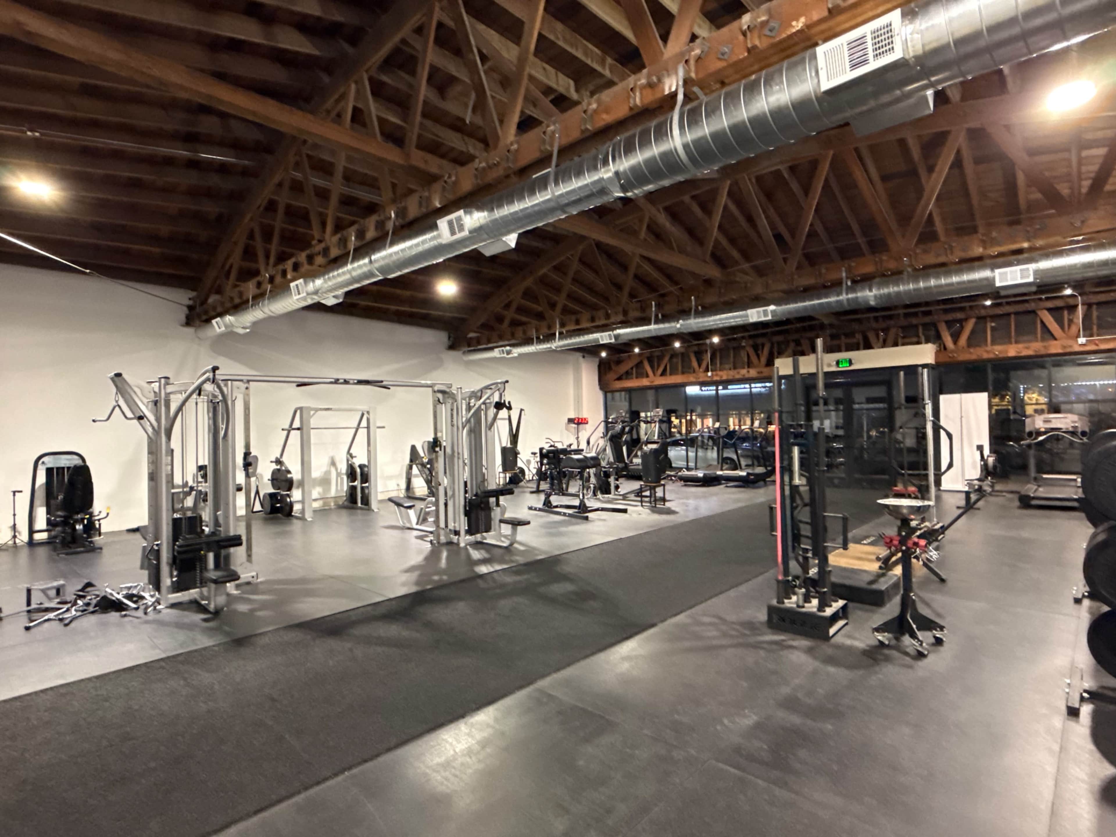 The image shows a modern gym interior with various exercise equipment, including weight machines, free weights, and workout stations, under a wooden beam ceiling.