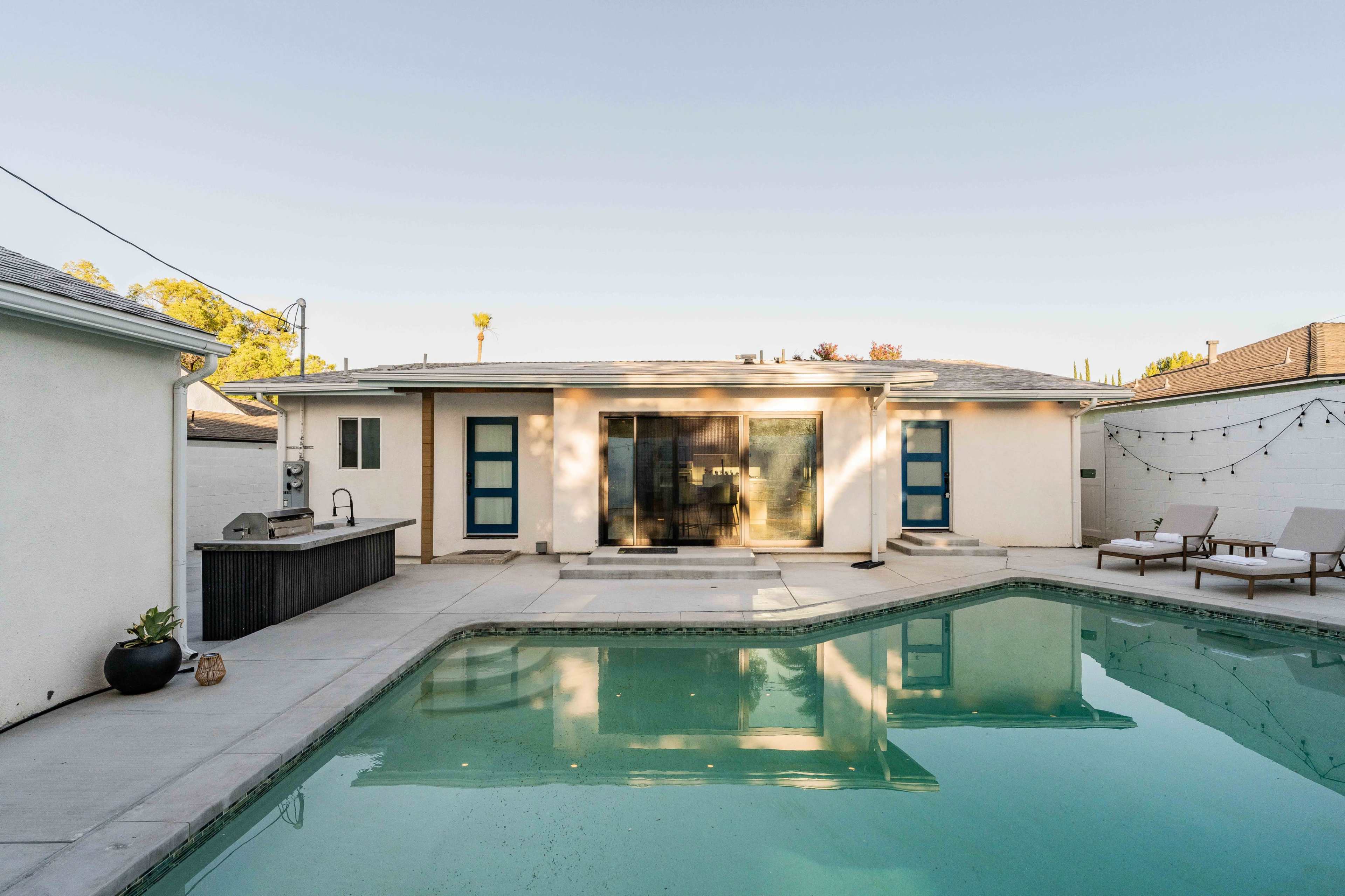 A swimming pool reflects a single-story house with blue doors and a barbecue area in the backyard.