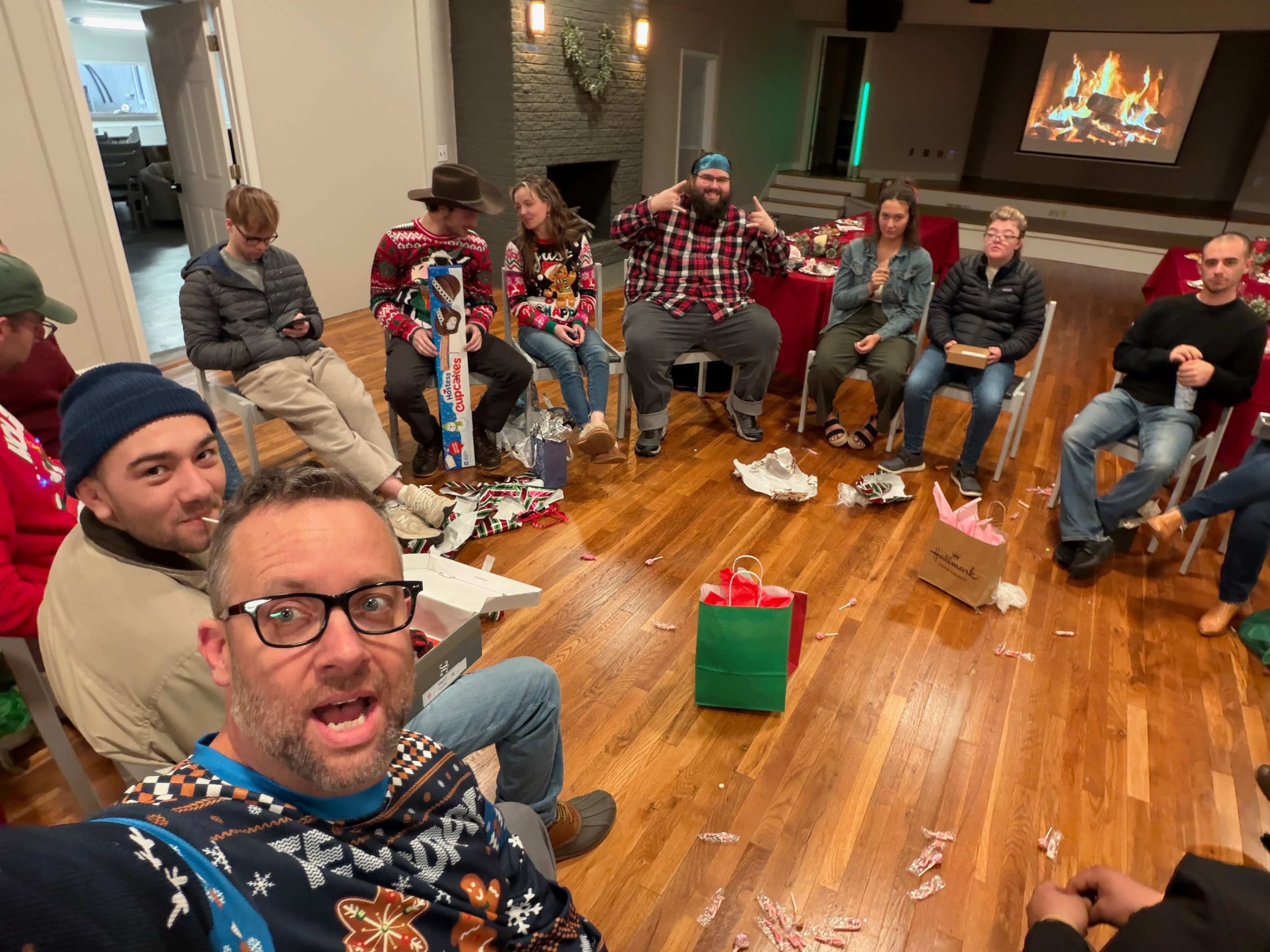 A group of people, some wearing festive sweaters, is seated in a circle on a wooden floor while wrapping paper and gifts are scattered around them.