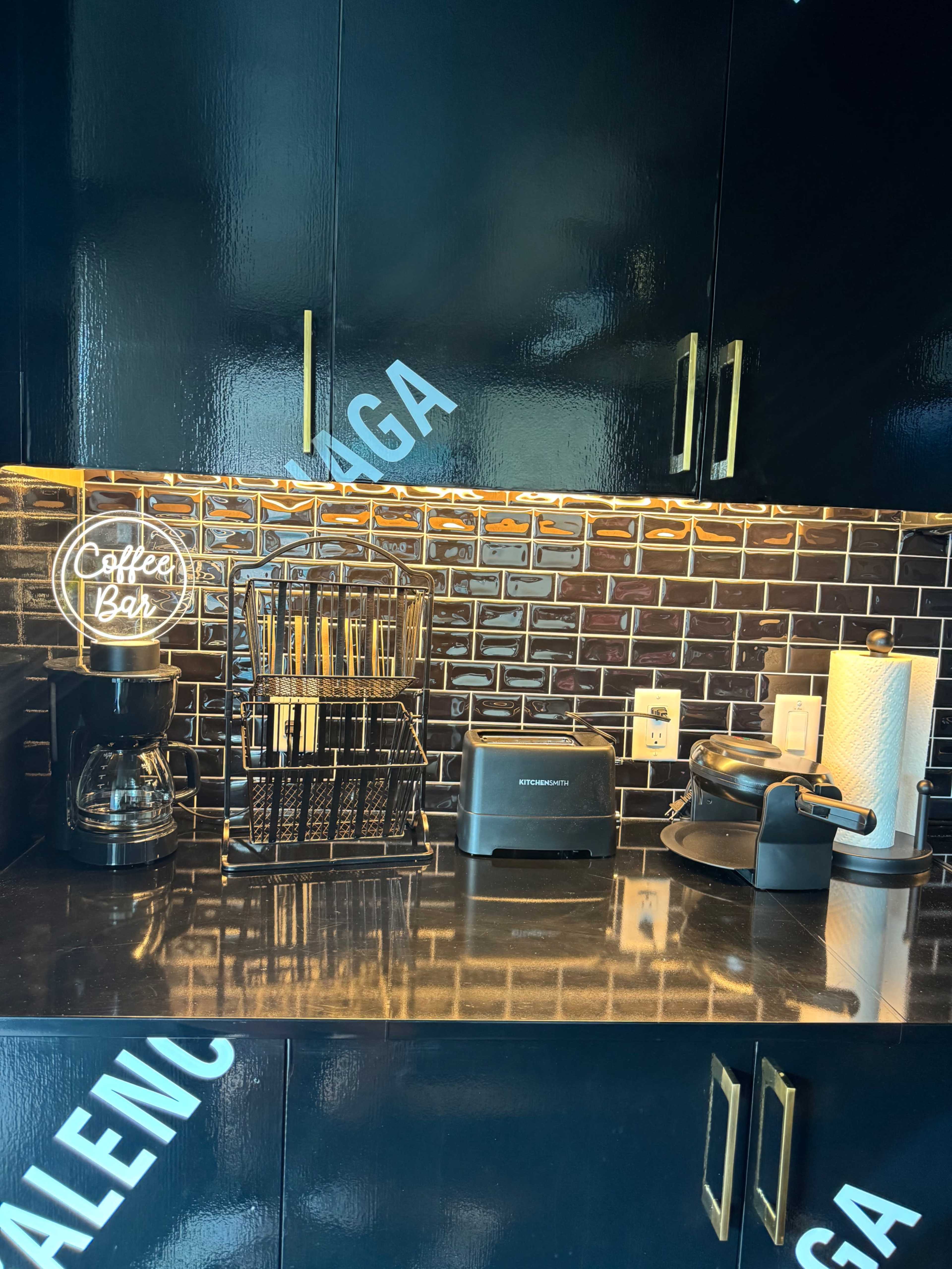 The image shows a kitchen counter with a coffee maker, toaster, and a waffle maker, along with a decorative "Coffee Bar" sign above a tiled backsplash.