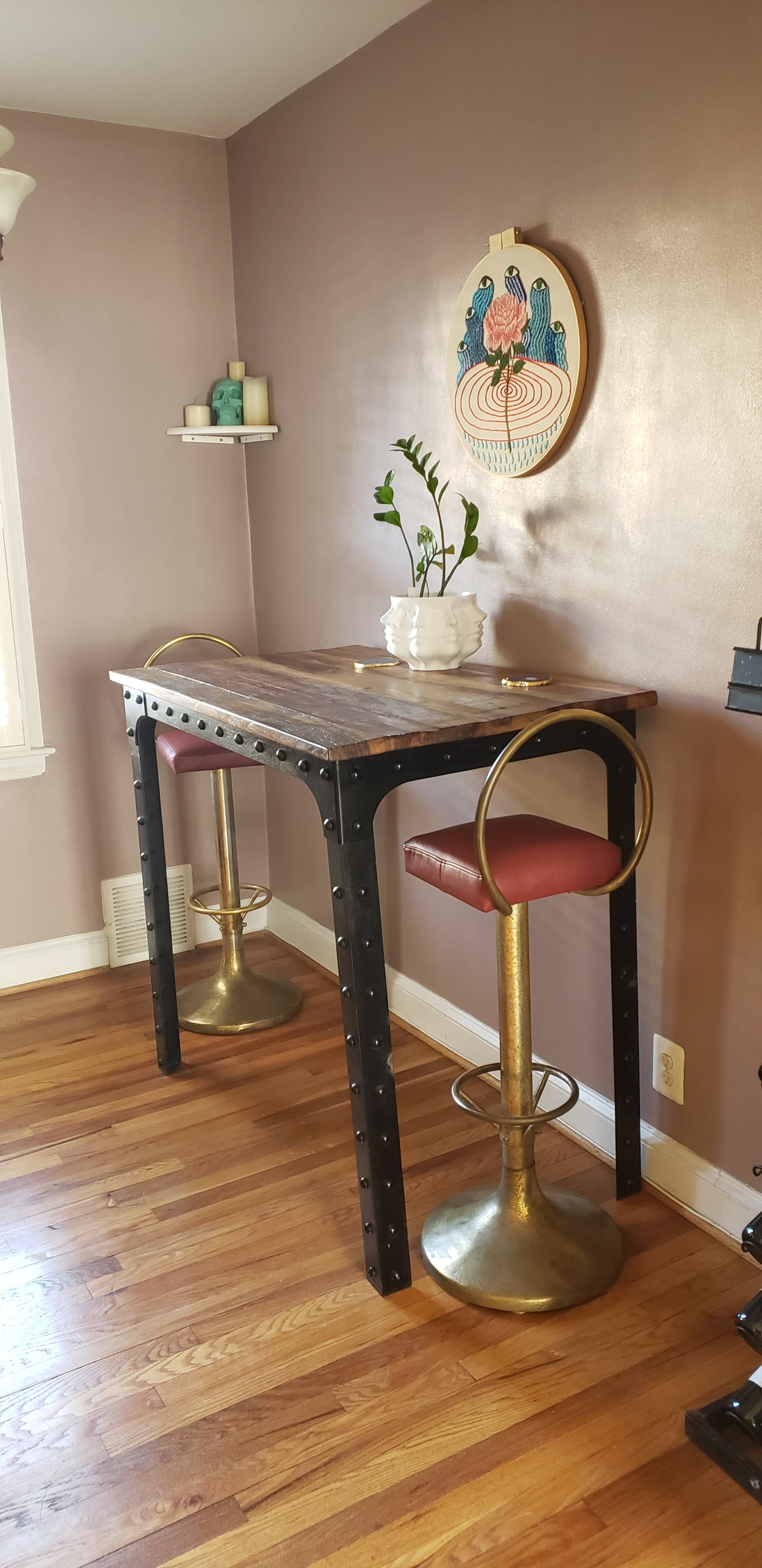 A wooden high table with black metal legs and two gold and red bar stools in a well-lit room adorned with a decorative wall piece and a small plant.