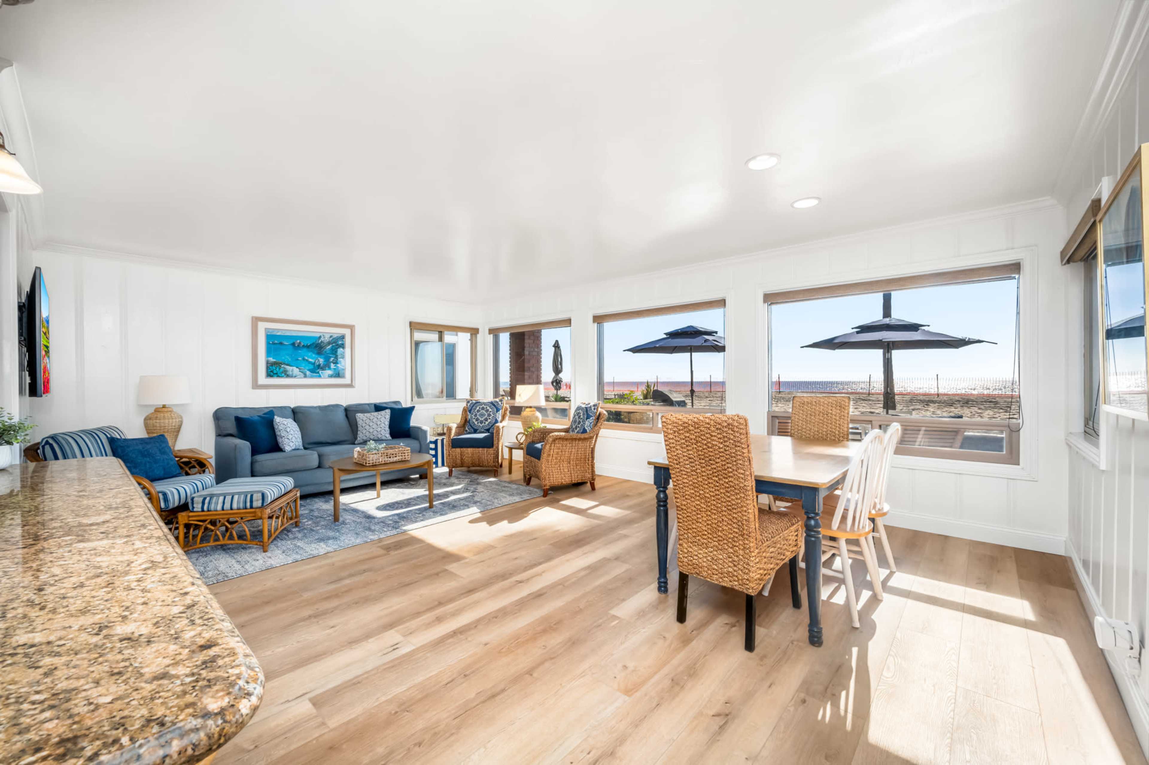 A bright, open living area with a dining table, a blue sofa, and large windows revealing a view of outdoor umbrellas.