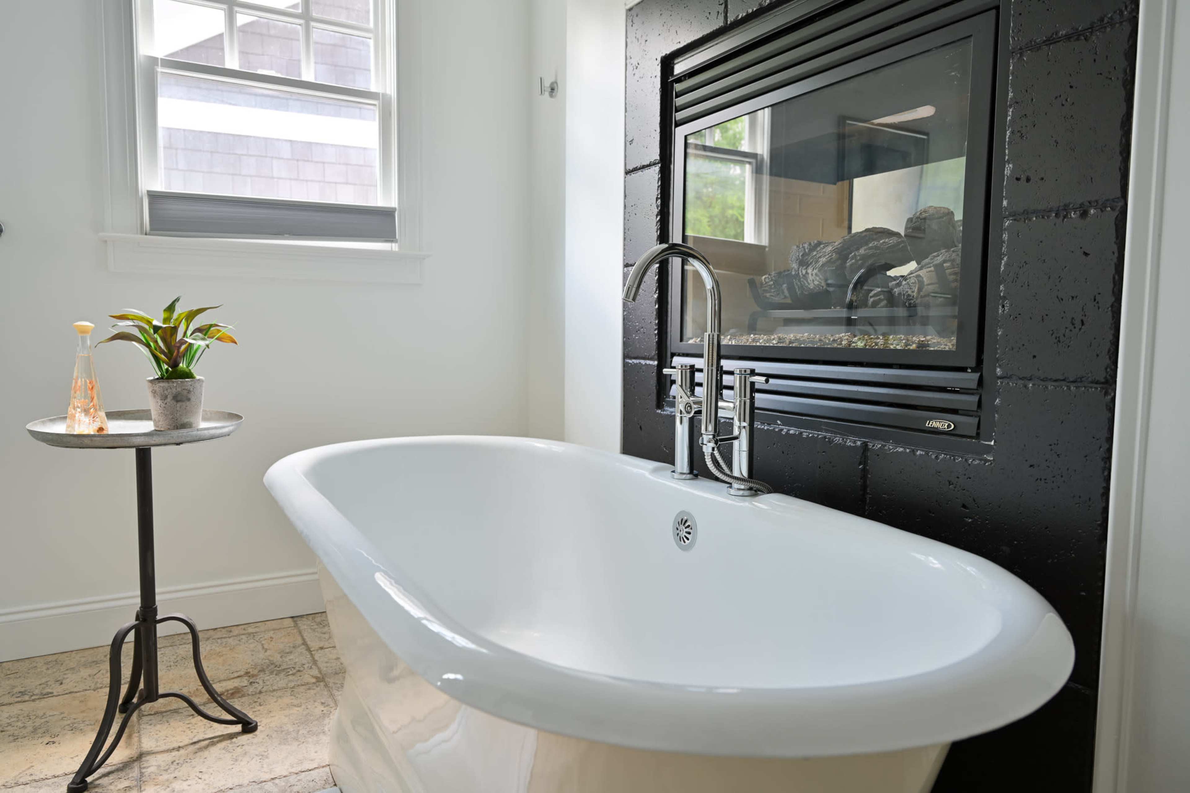 A modern bathroom with a freestanding white bathtub, a small table with a potted plant, and a fireplace embedded in a feature wall.