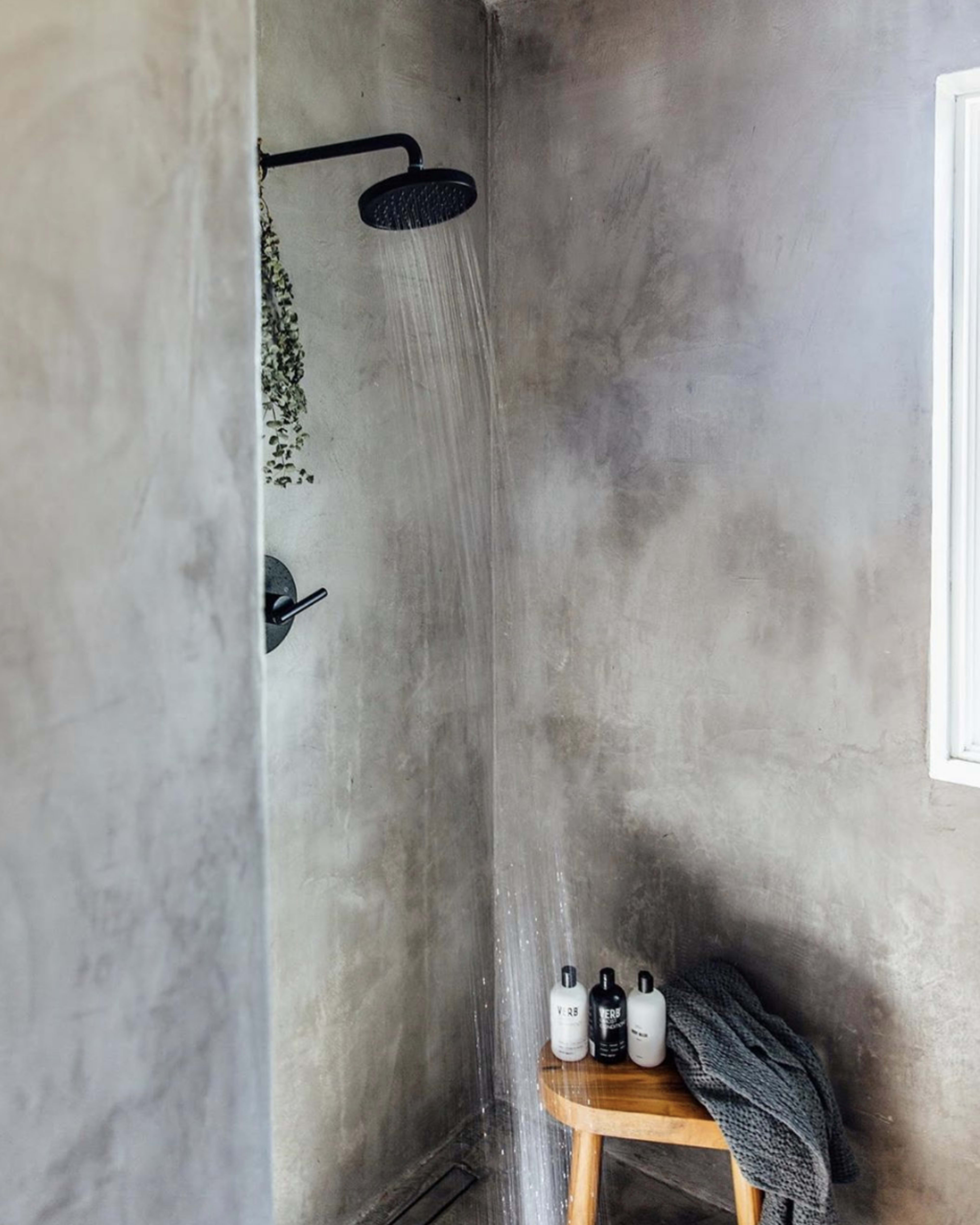 The image shows a minimalist shower area with a rain showerhead, concrete walls, and a wooden stool holding three bottles and a grey towel.