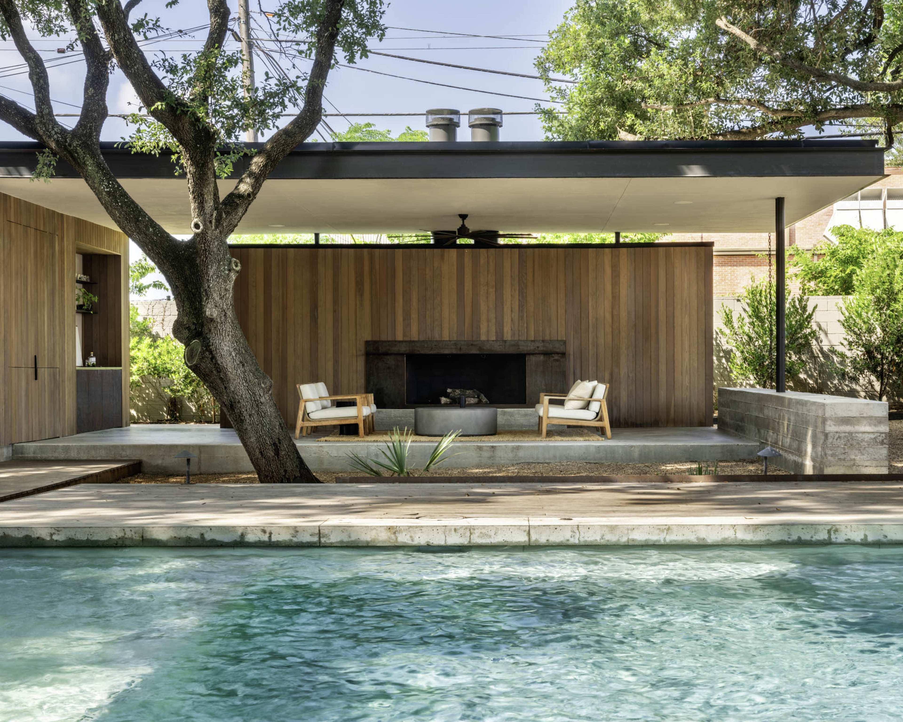 A modern outdoor space features a pool, two chairs positioned by a fireplace, and a wooden structure surrounded by greenery.