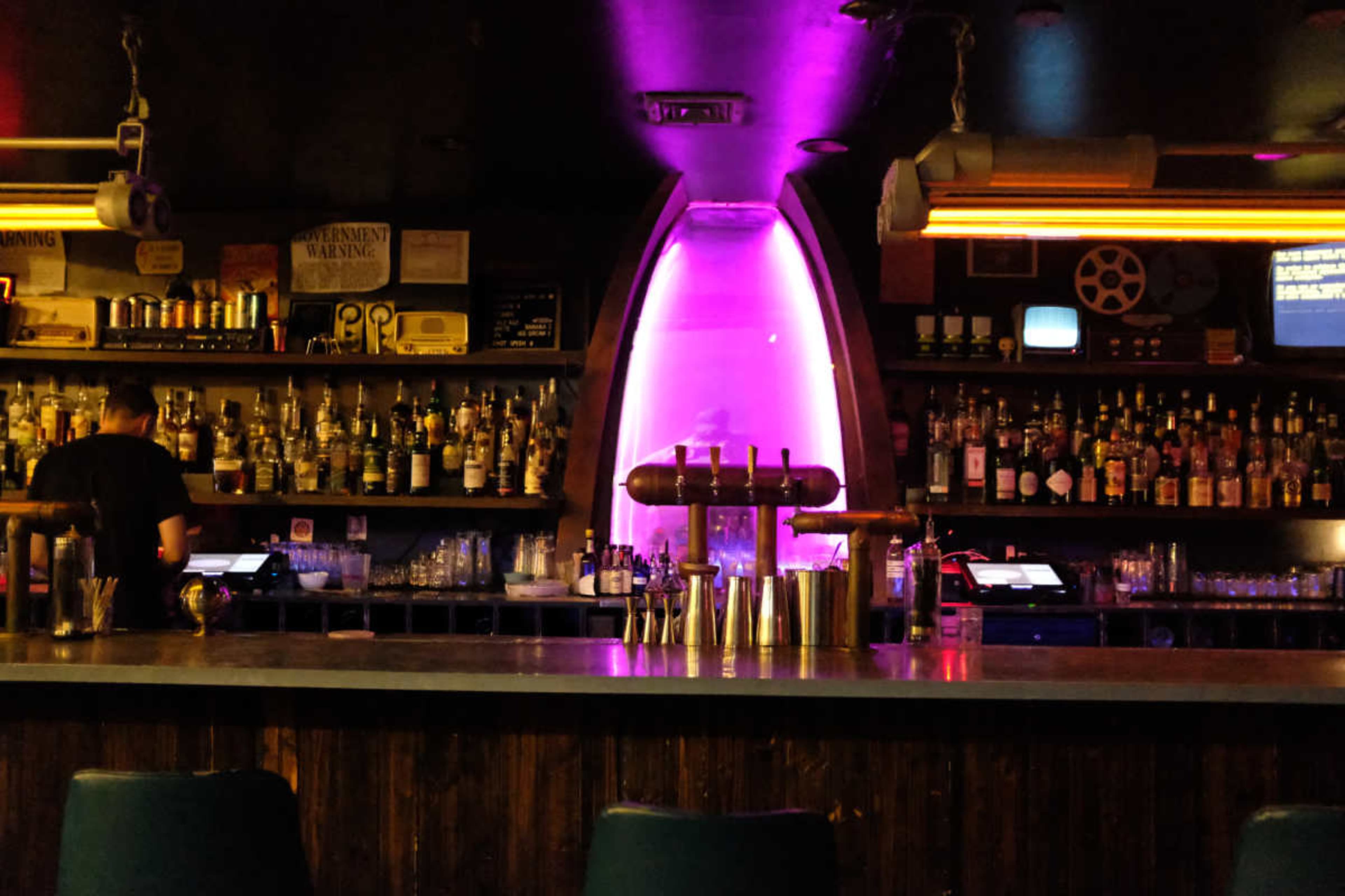 A dimly lit bar features a well-stocked shelf of bottles behind the counter, with a bartender preparing drinks in the foreground.