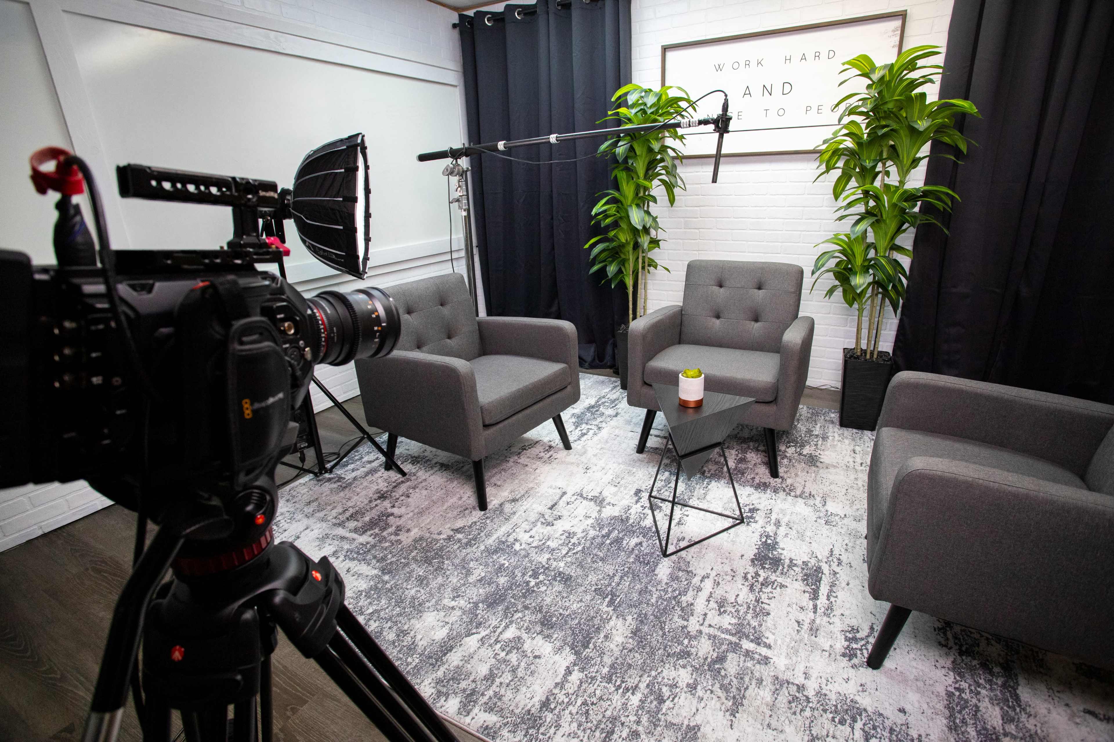 A professional video setup features three gray armchairs arranged on a rug, a small table with a drink, and potted plants, with a camera and lighting equipment ready for recording.