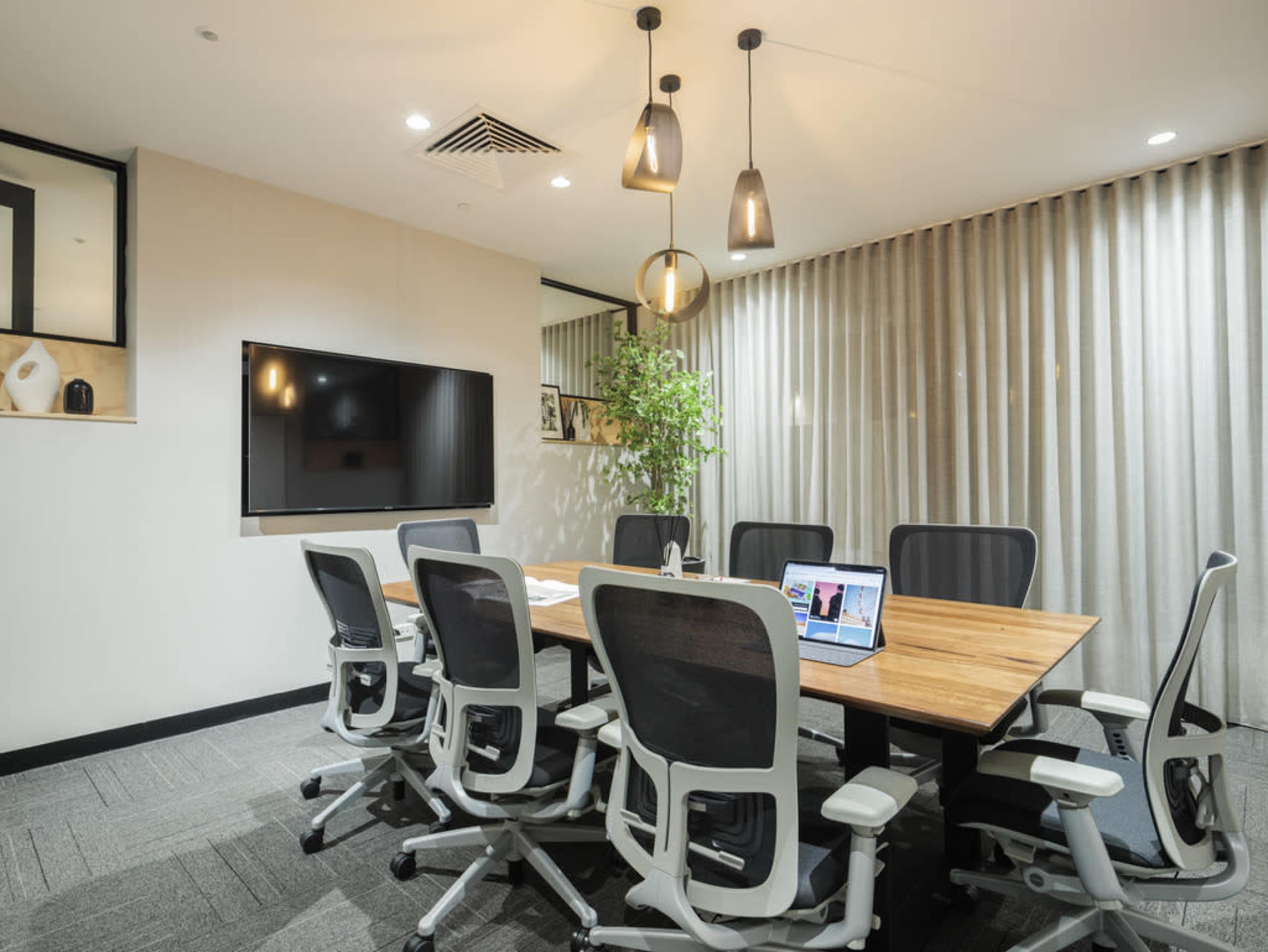 A modern conference room with a large wooden table, six ergonomic chairs, a wall-mounted television, and a potted plant, illuminated by pendant lights.