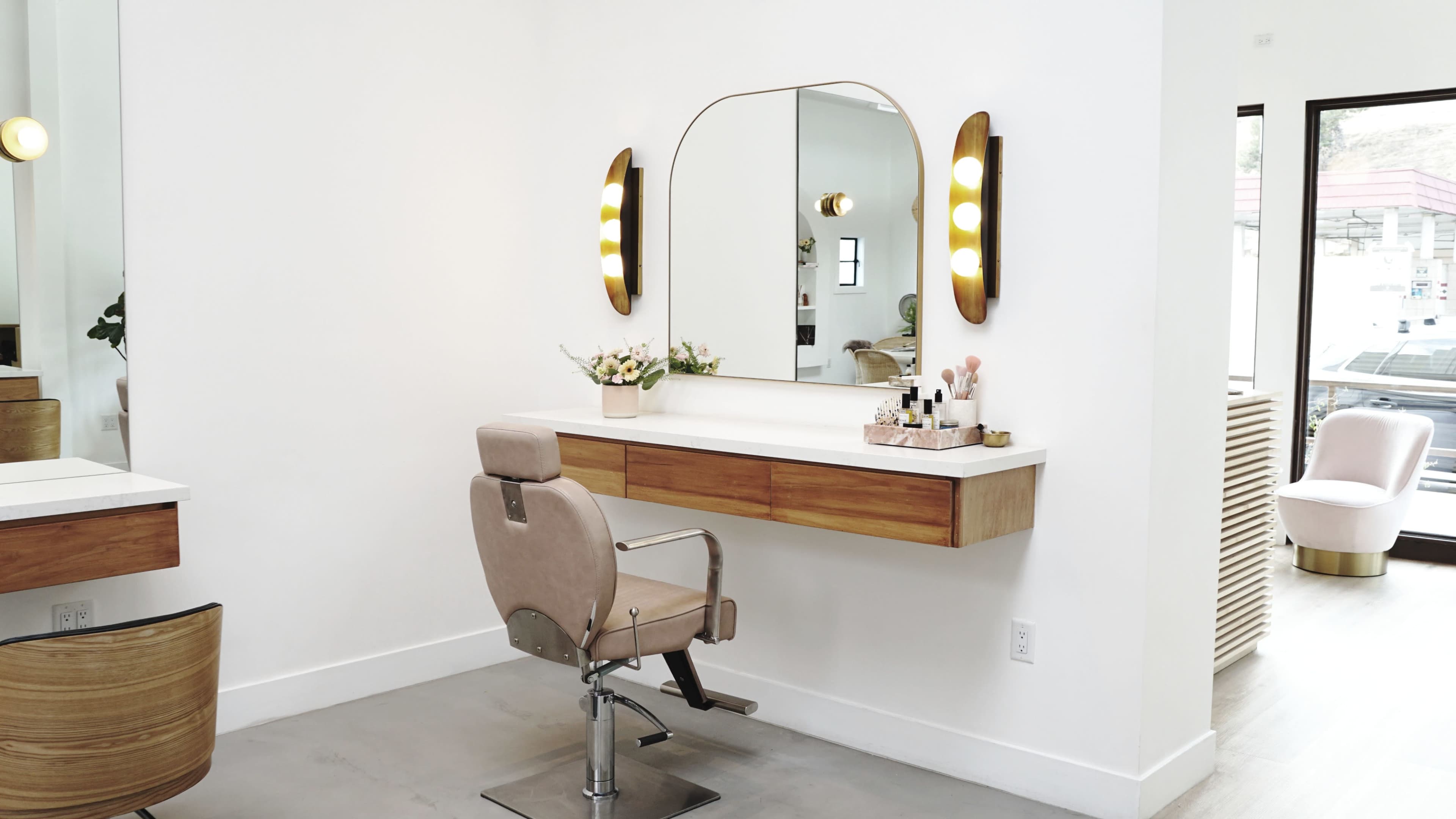 The image shows a modern hair salon interior with a wooden vanity, a large mirror surrounded by rounded light fixtures, and a beige styling chair.