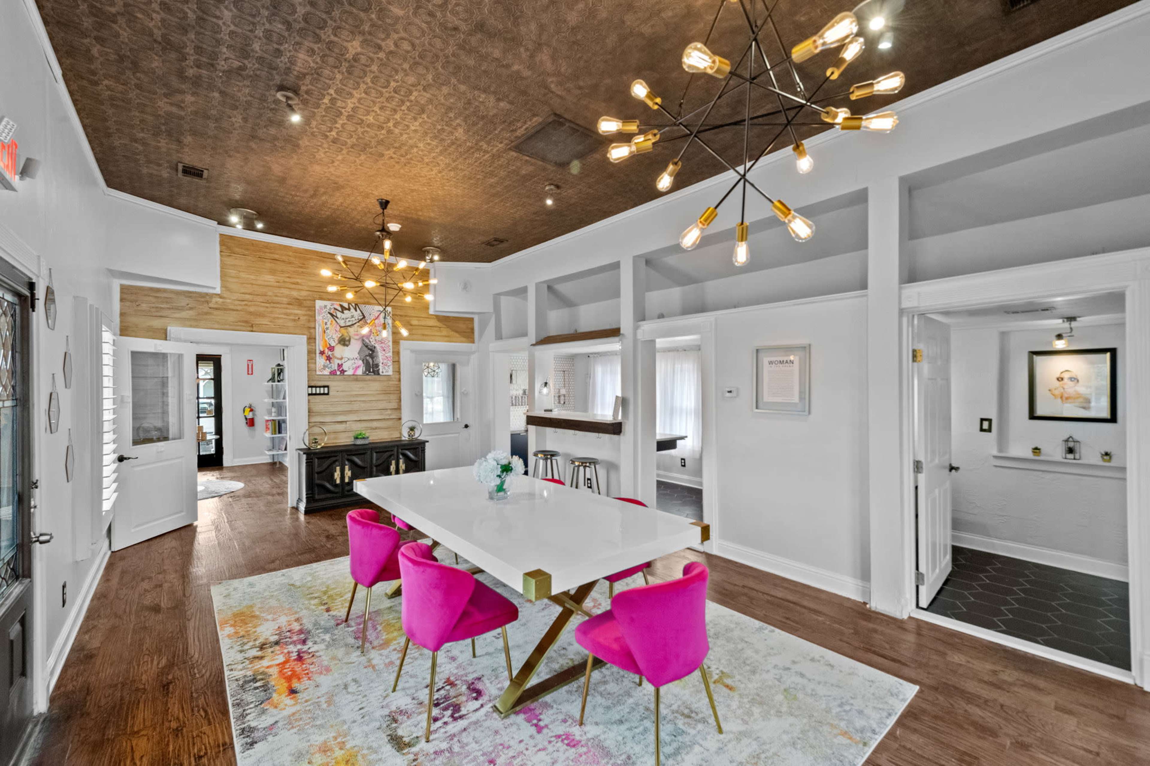 A modern interior space features a dining table surrounded by pink chairs, sleek lighting fixtures, and wooden accents on the walls.