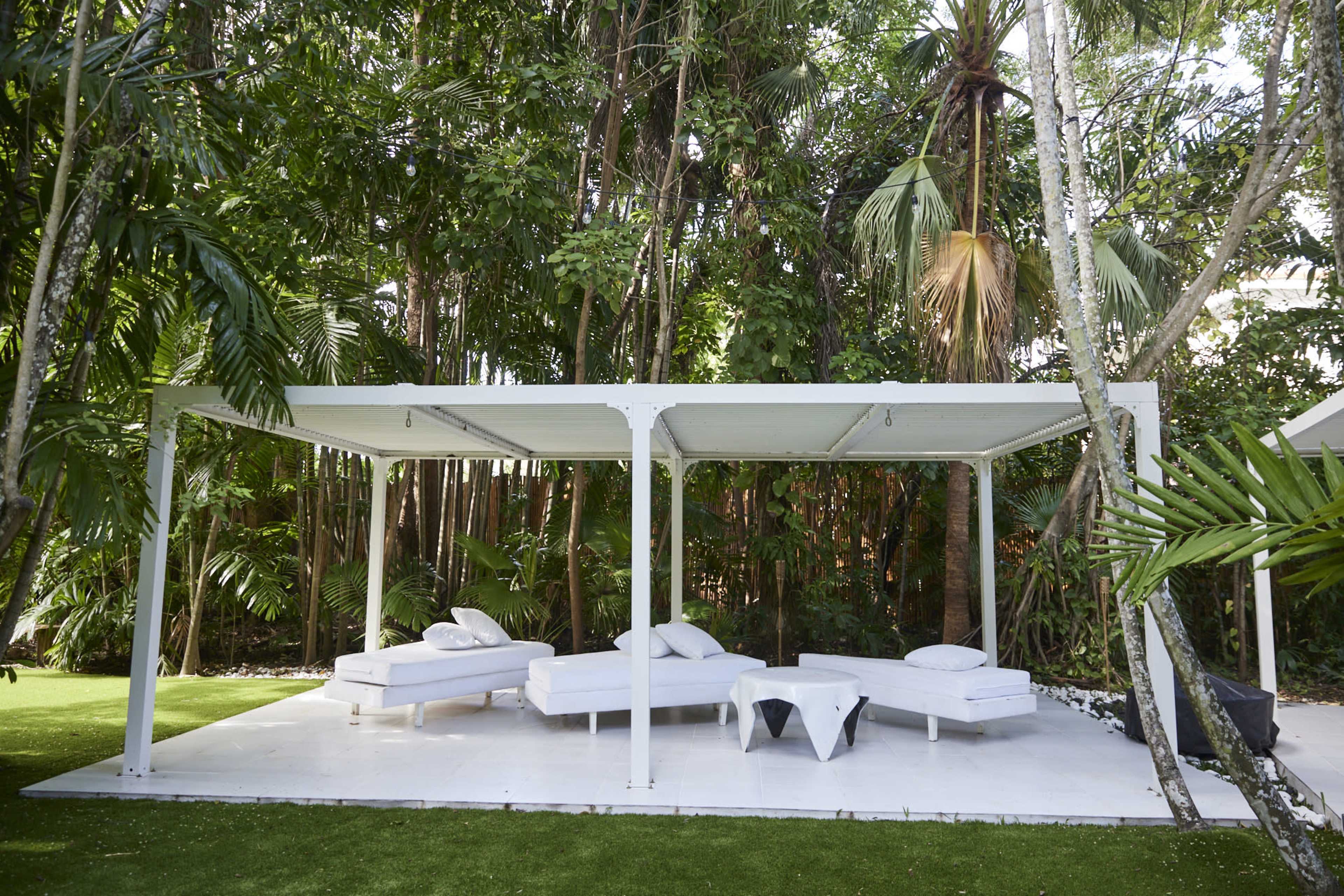 A white outdoor lounge area with several beds is situated under a pergola, surrounded by lush tropical vegetation.