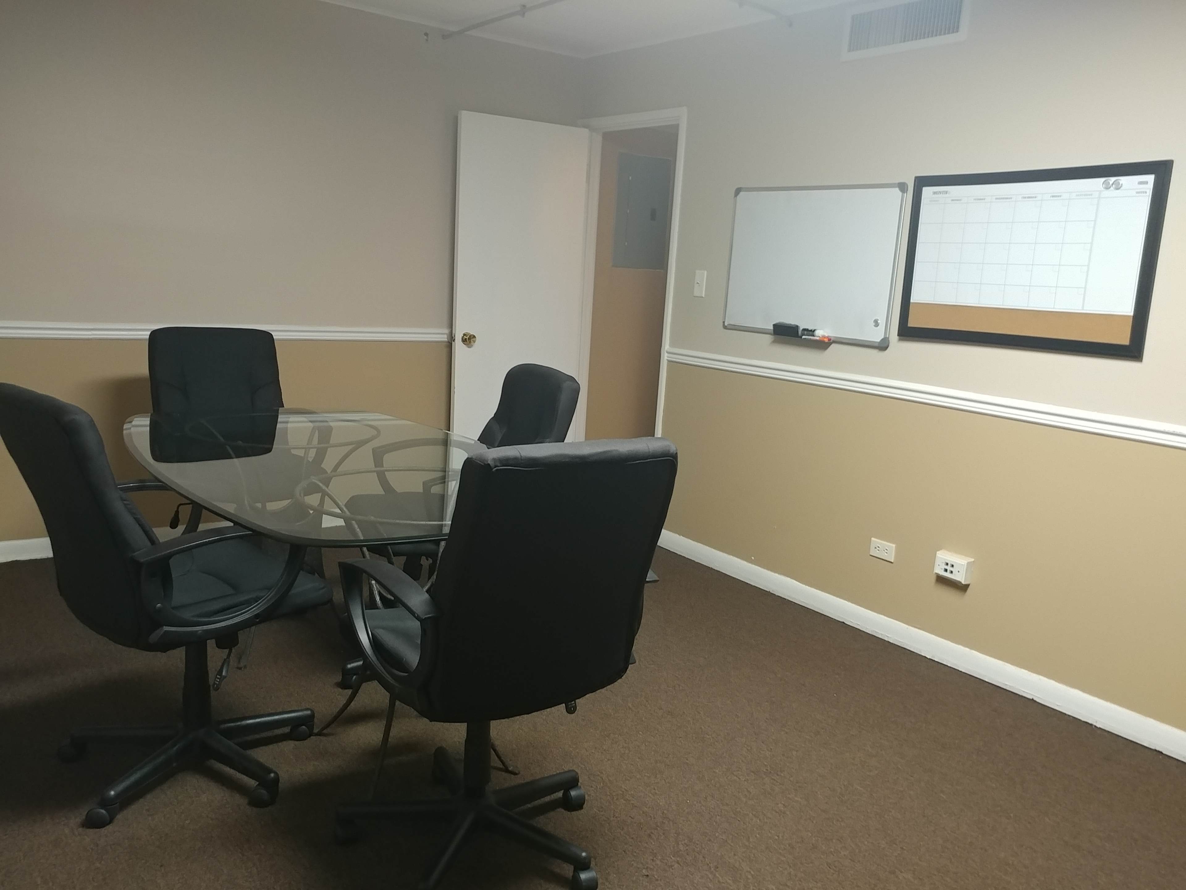 The image shows a small conference room with a glass table, four black chairs, a whiteboard mounted on the wall, and a calendar framed beside it.