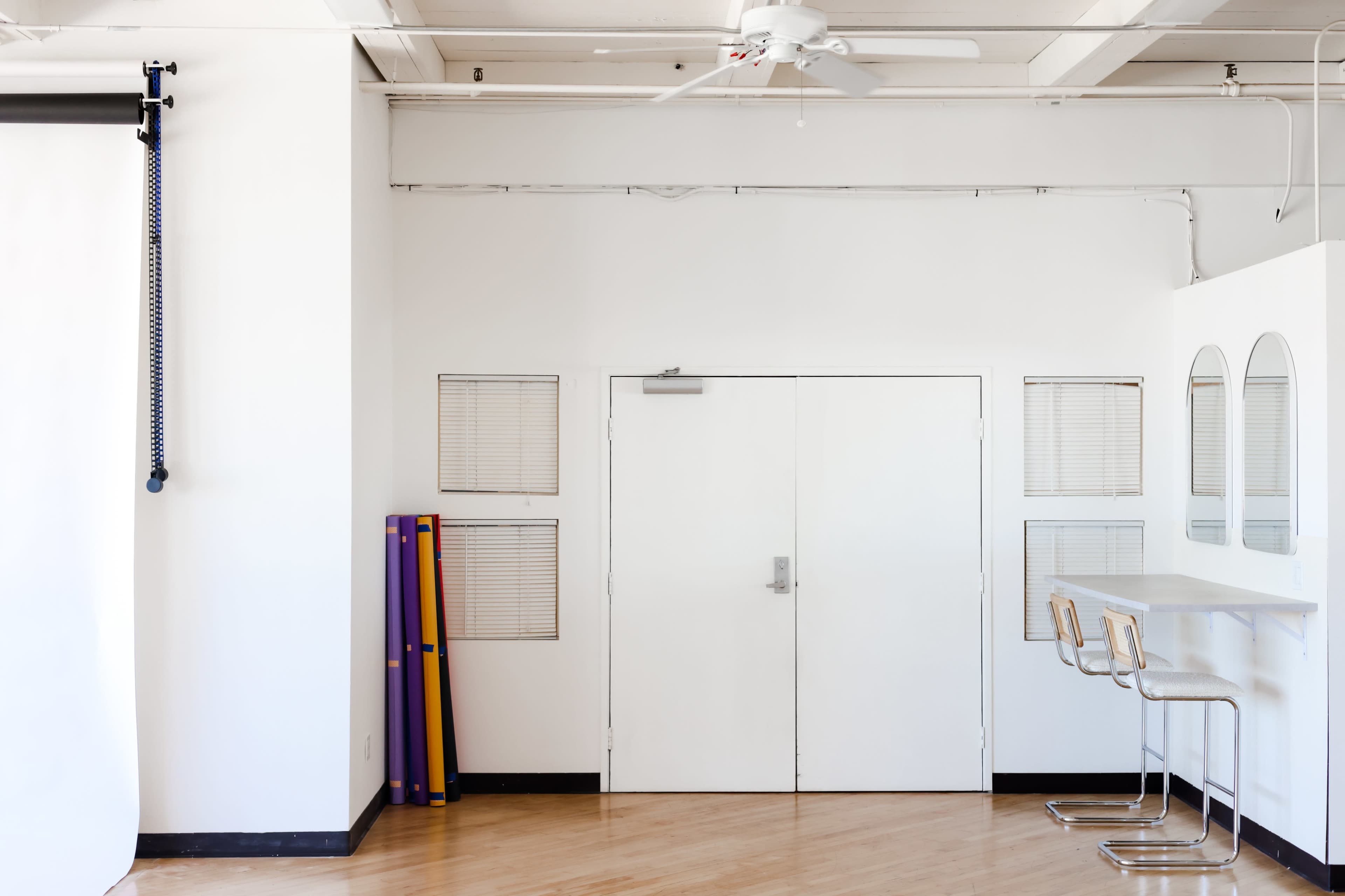 A minimalistic studio space with white walls, a closed door, two round mirrors, and three barstools beside a countertop.