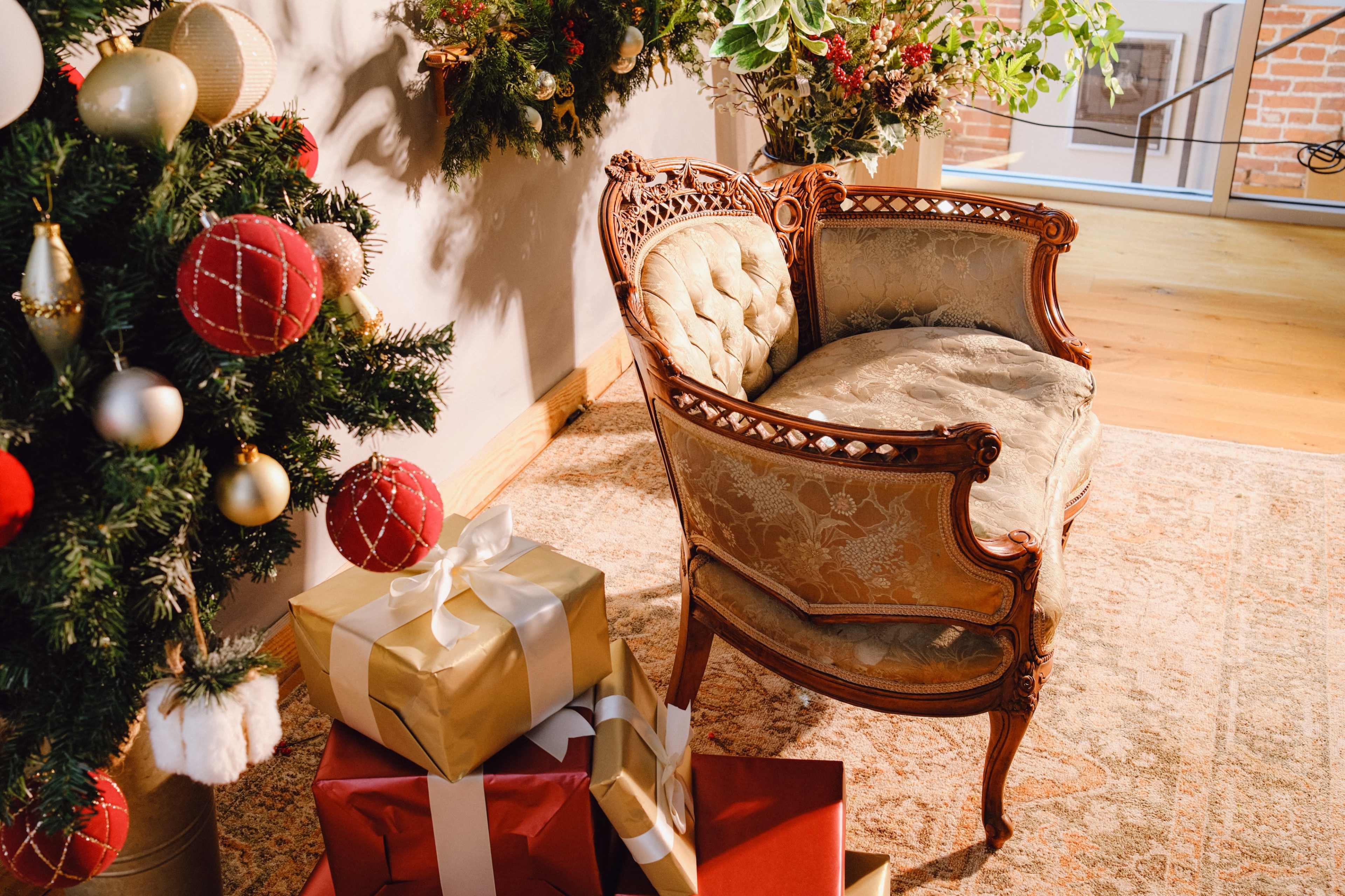 A vintage upholstered chair is positioned next to a Christmas tree adorned with ornaments, with several wrapped gifts stacked nearby on a patterned rug.