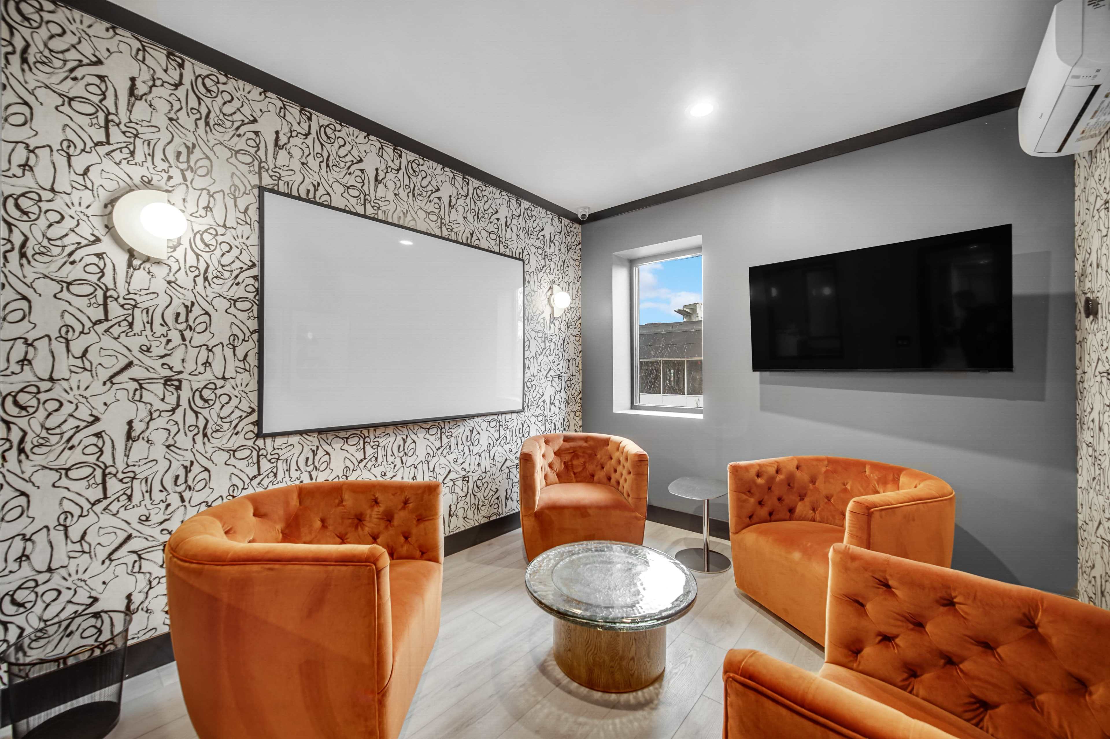 A small meeting room features orange armchairs, a round glass table, and a large wall-mounted television, with decorative wallpaper and a bright window.