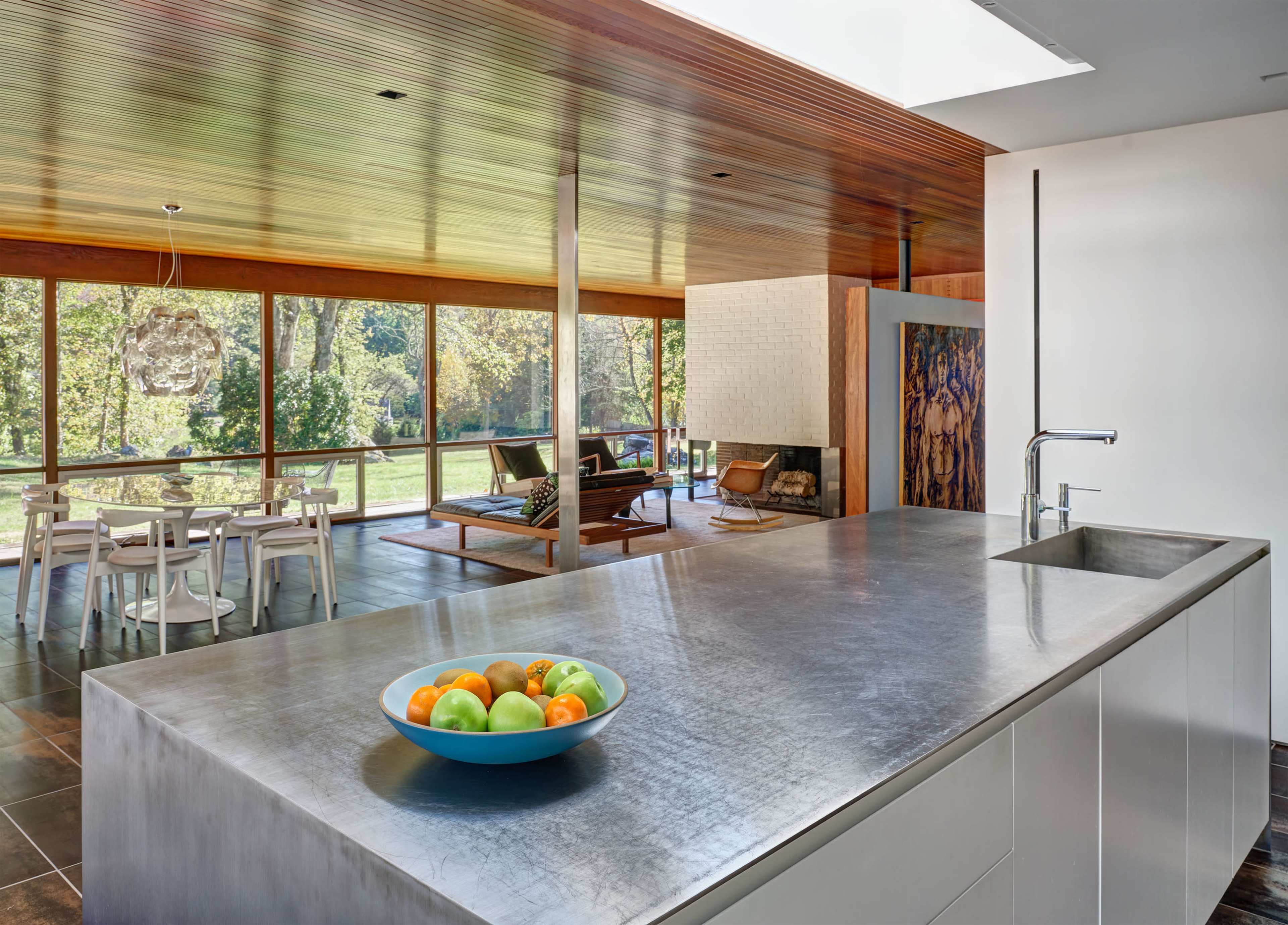 The kitchen features a stainless steel island with a bowl of fruit, overlooking a spacious dining area and large windows that reveal a view of the outdoor landscape.