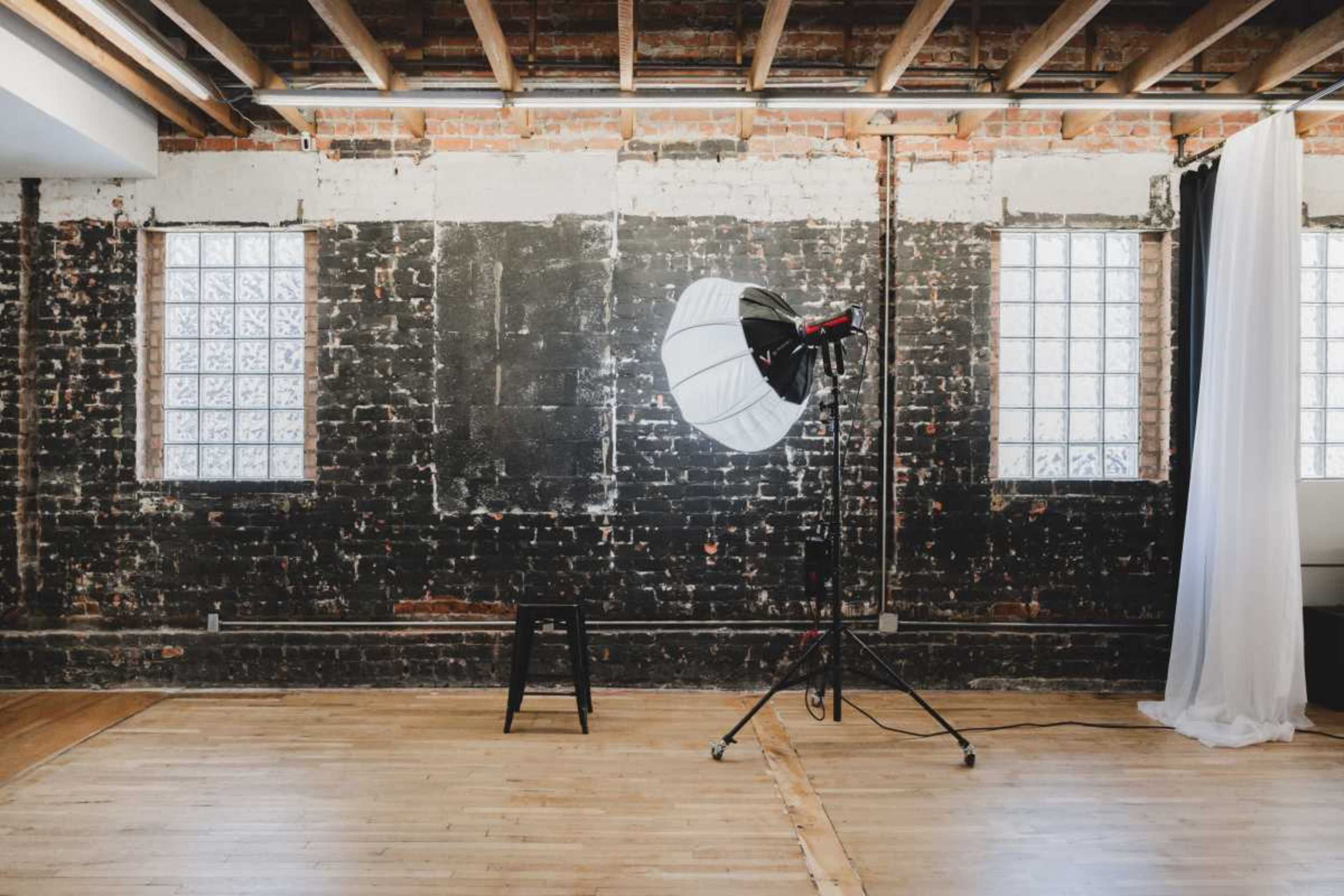 A photography studio features a large softbox on a stand, a stool, and exposed brick walls with windows.