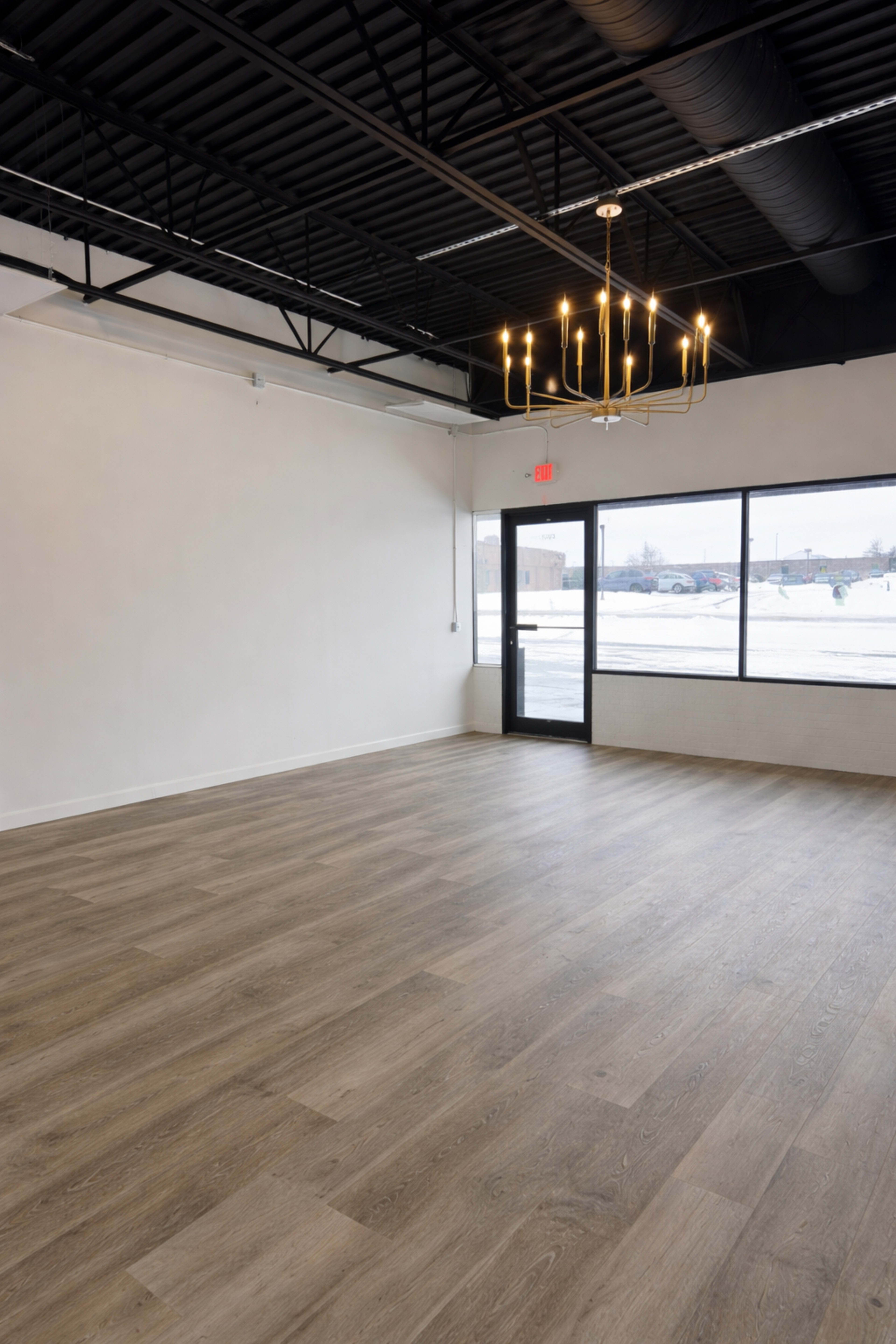 The image shows an empty room with light wood flooring, white walls, and a modern chandelier, featuring large windows displaying a snowy outdoor scene.