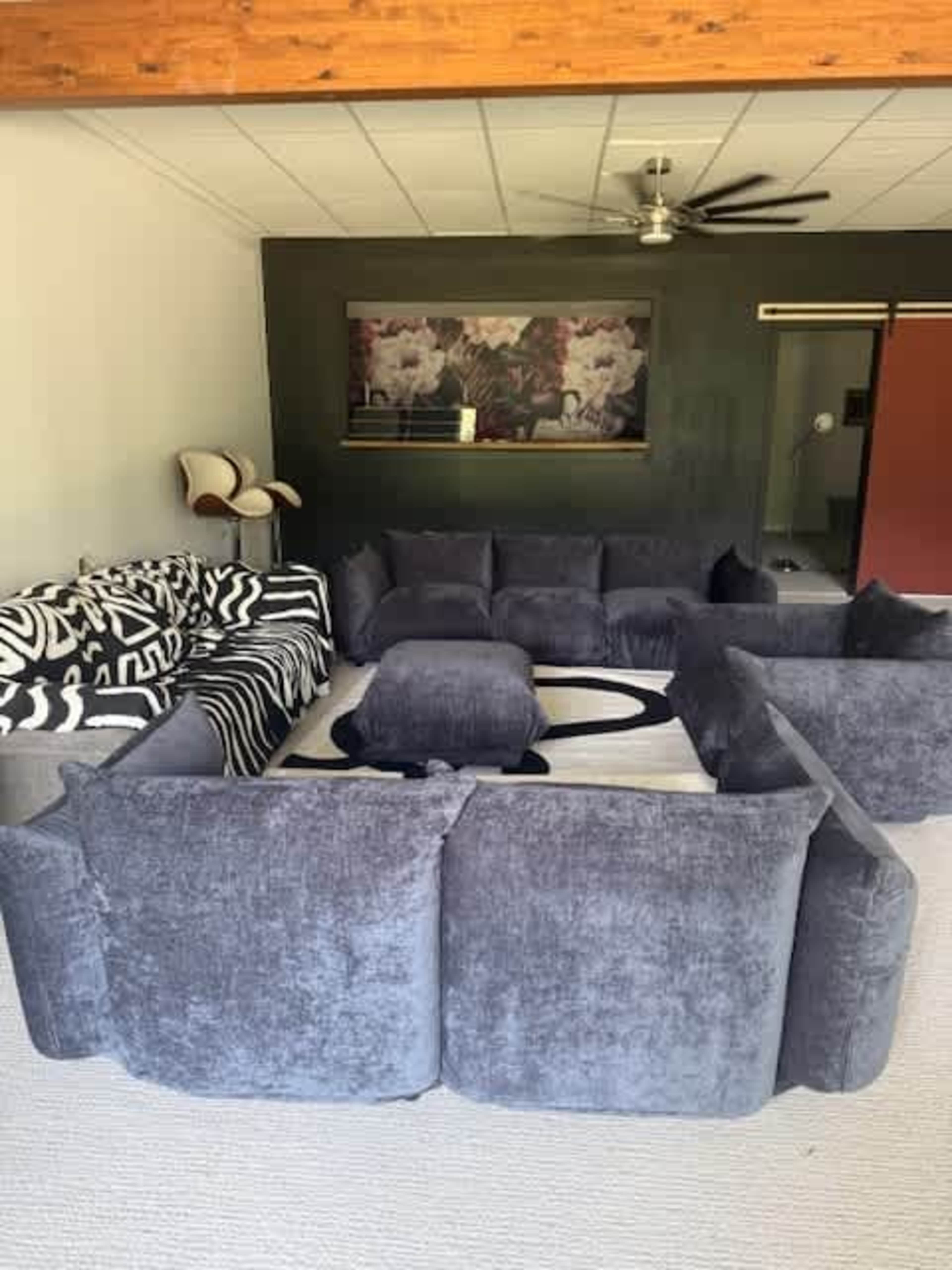 The room features a seating arrangement made up of multiple gray fabric sofas arranged in a circular formation, with a coffee table in the center and a large artwork on the wall.