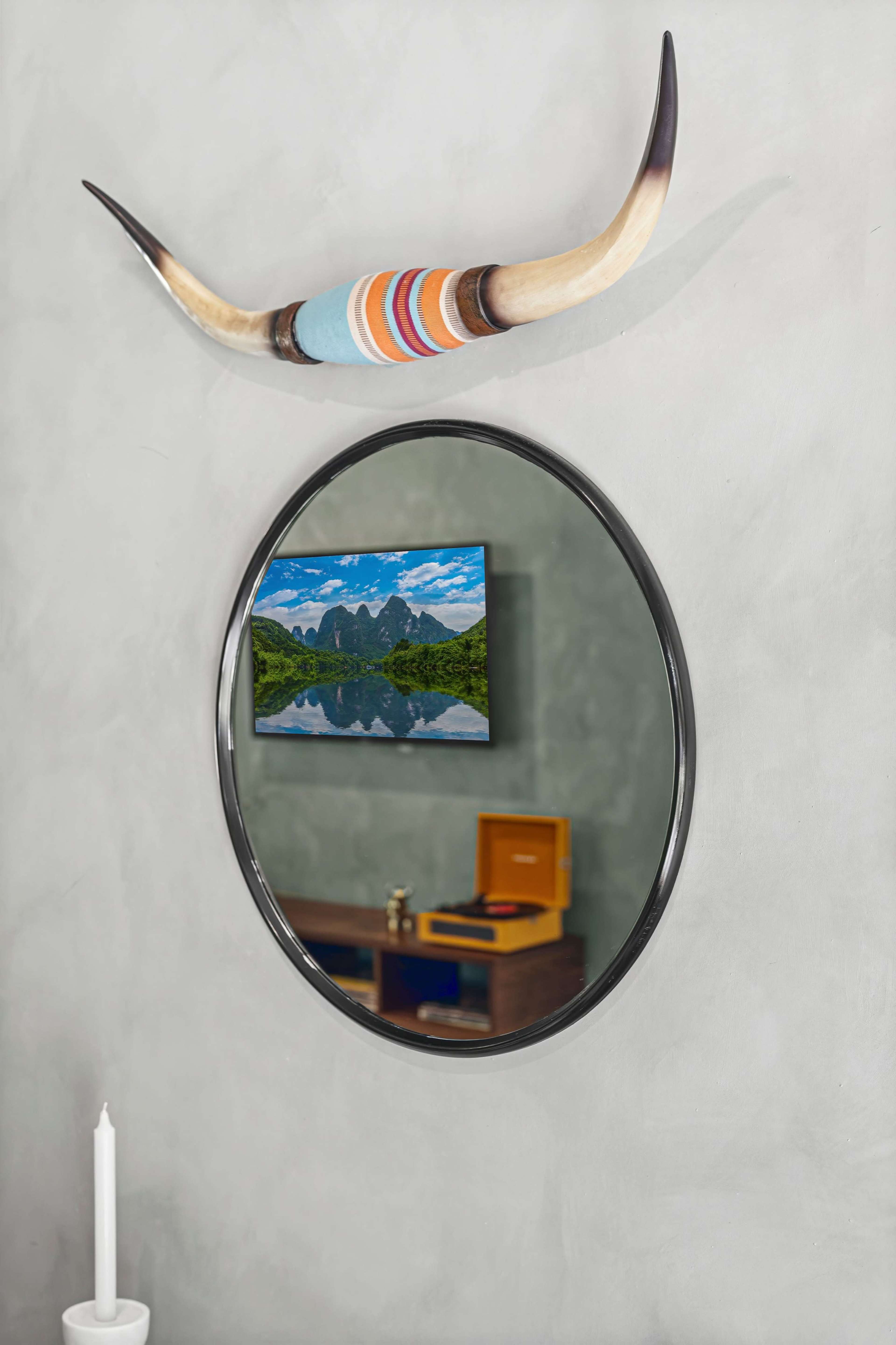 A round mirror hangs on a gray wall, reflecting a scenic landscape with mountains and a lake, alongside decorative horns above it and a wooden cabinet below.