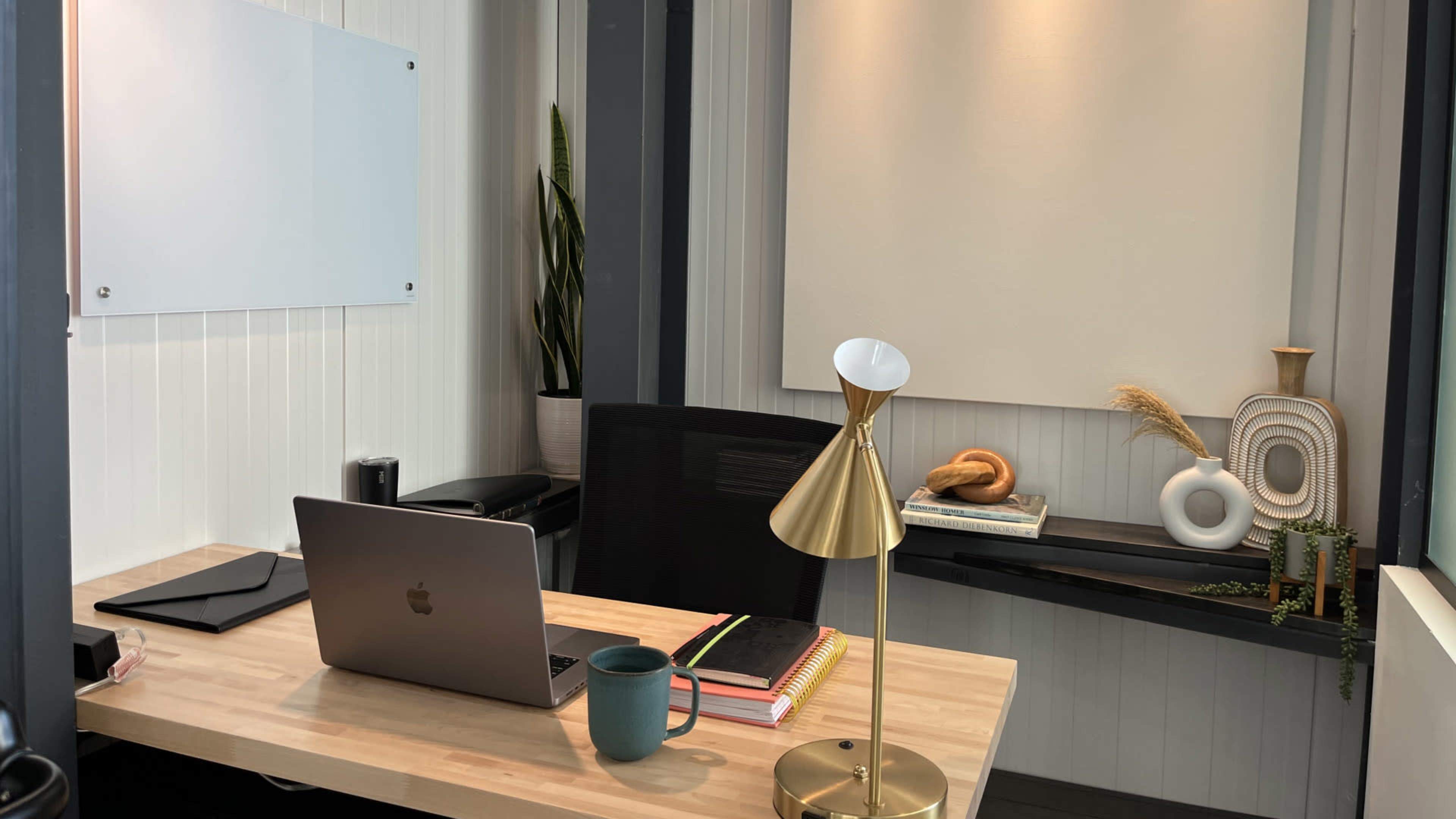 A modern office space features a wooden desk with a laptop, a coffee mug, books, a gold desk lamp, and decorative items on the wall shelf.