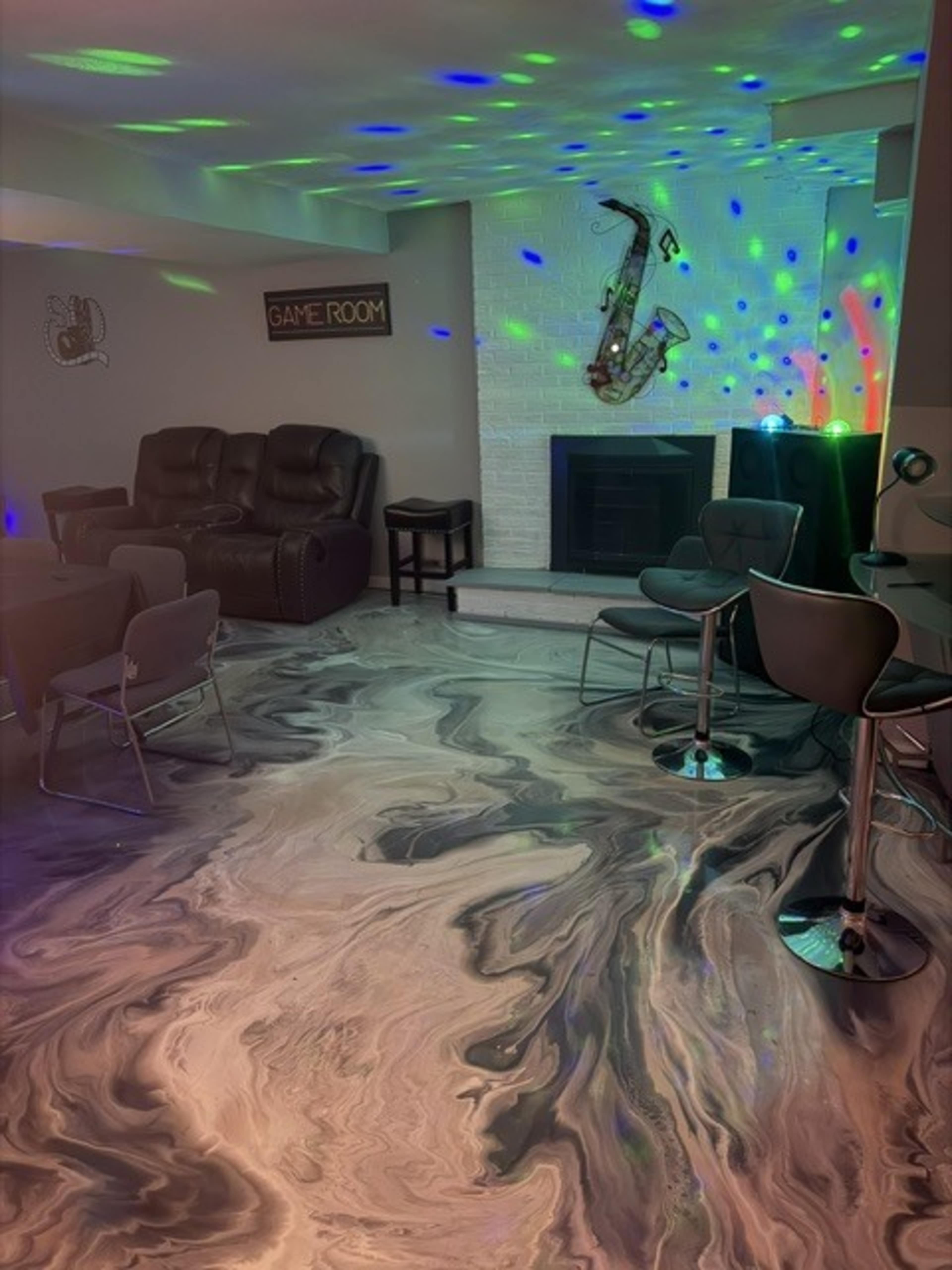 The image shows a game room with a marbled floor design, a fireplace, and seating arrangements that include recliners and bar stools, complemented by colorful lighting effects.
