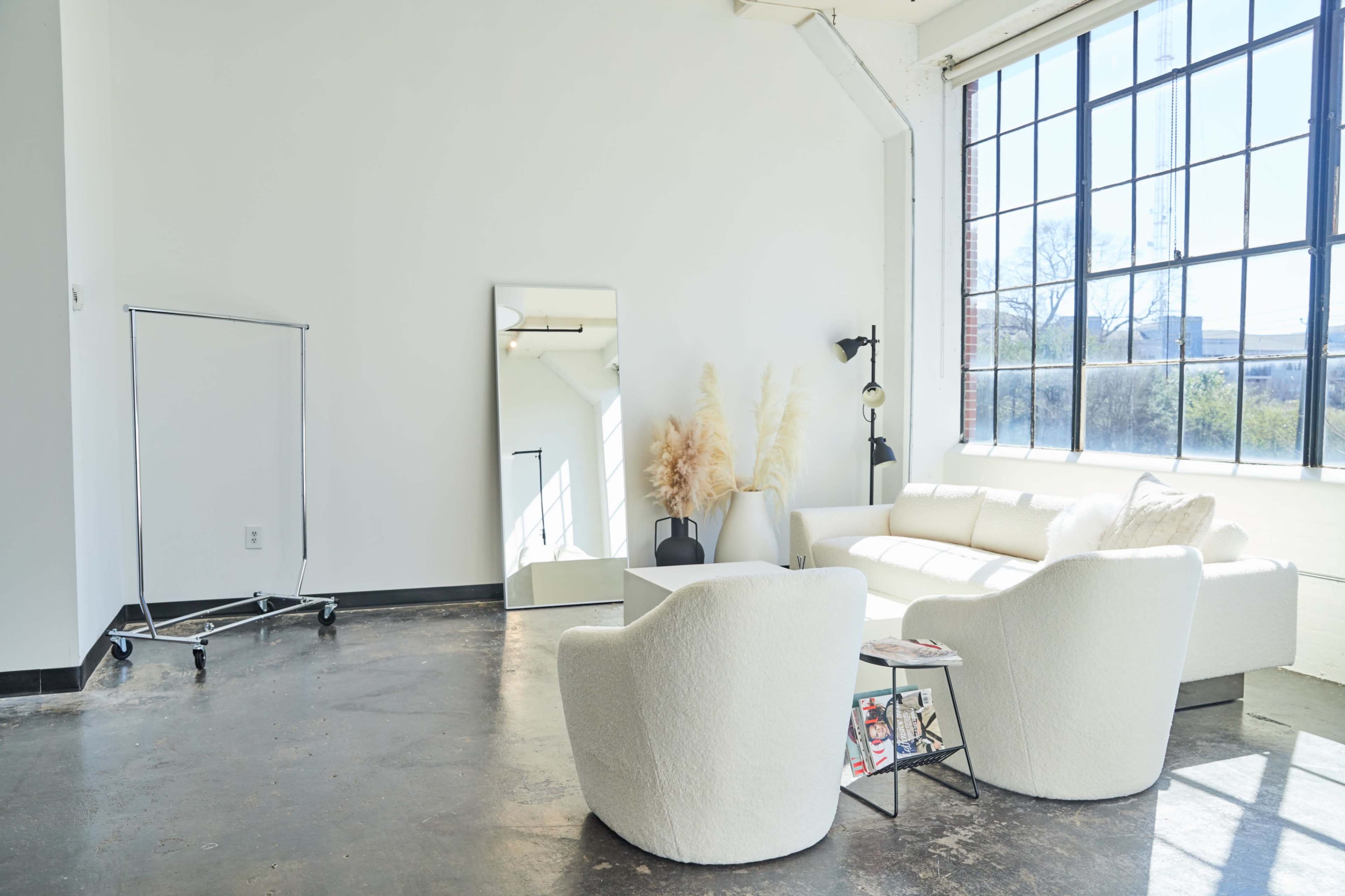 A minimalist living space featuring a white sofa, two round chairs, a large mirror, and a clothing rack, all set against a background of a large window.