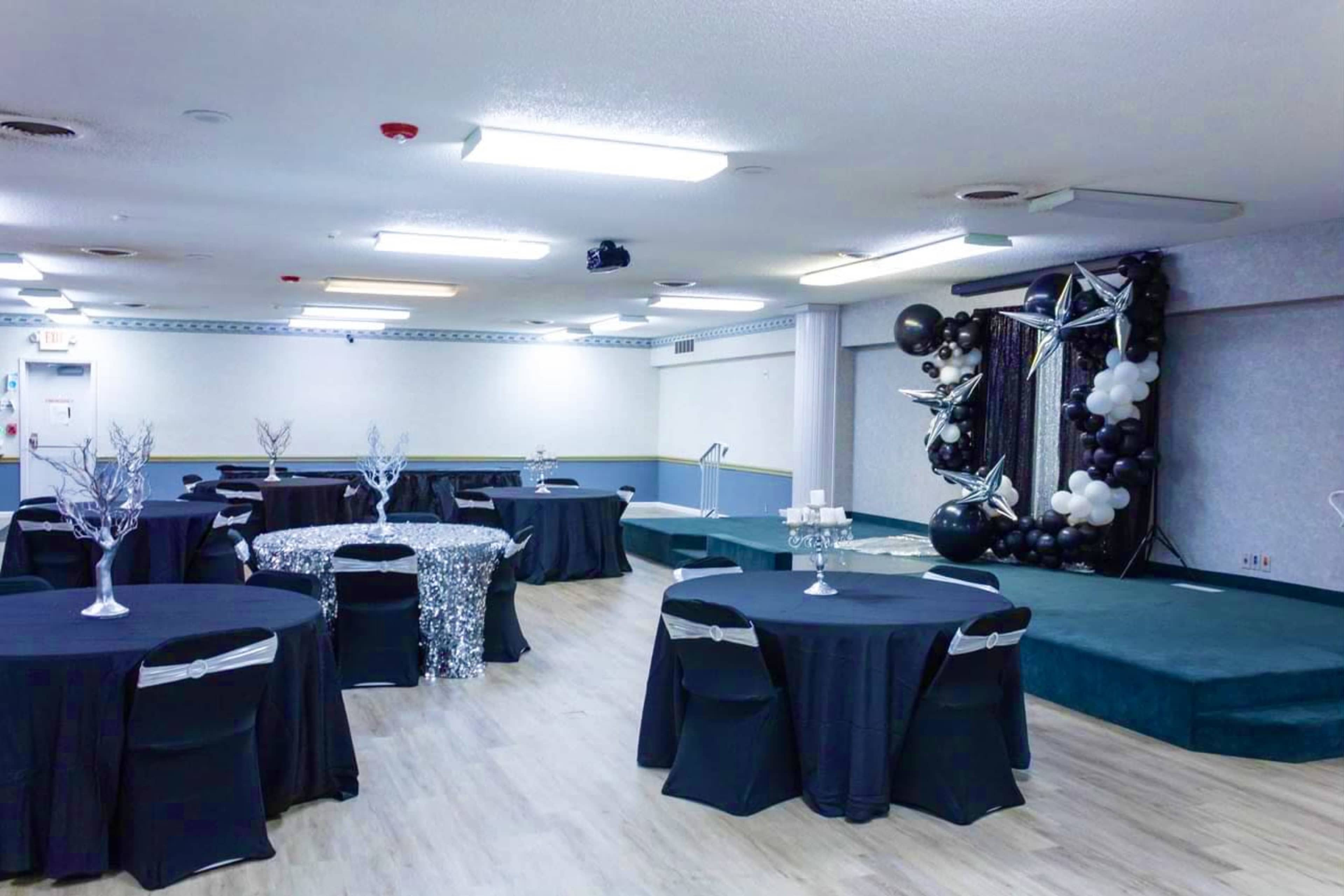 A large event space with black tables and a stage decorated with silver and black balloons.