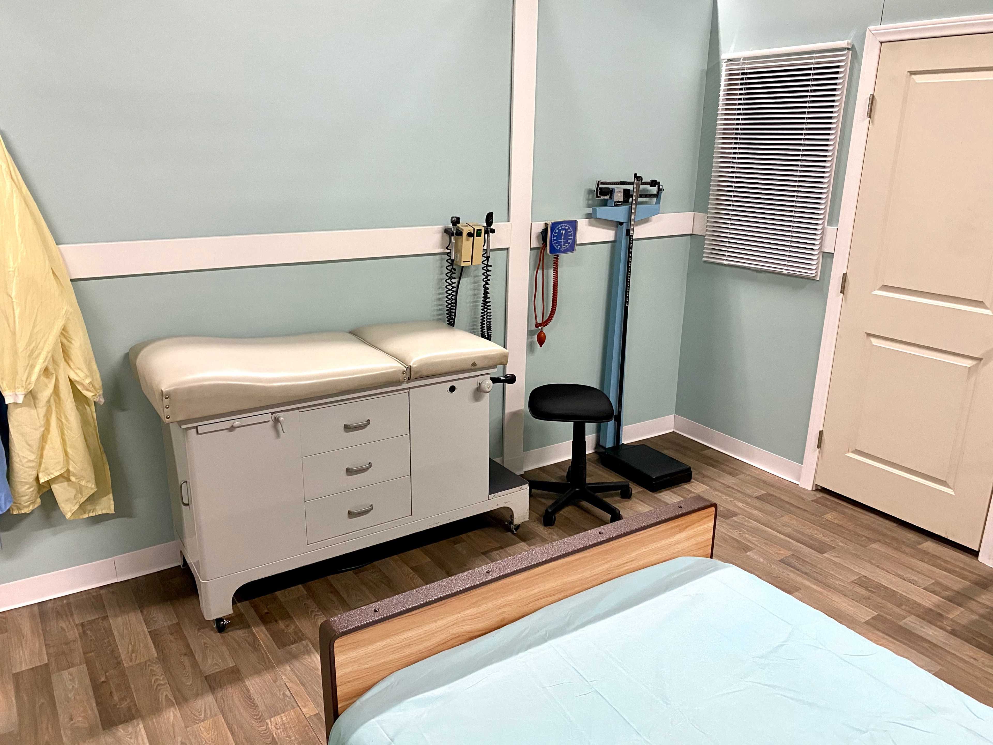 A medical examination room with an examination table, a chair, a weighing scale, and medical equipment on the wall.