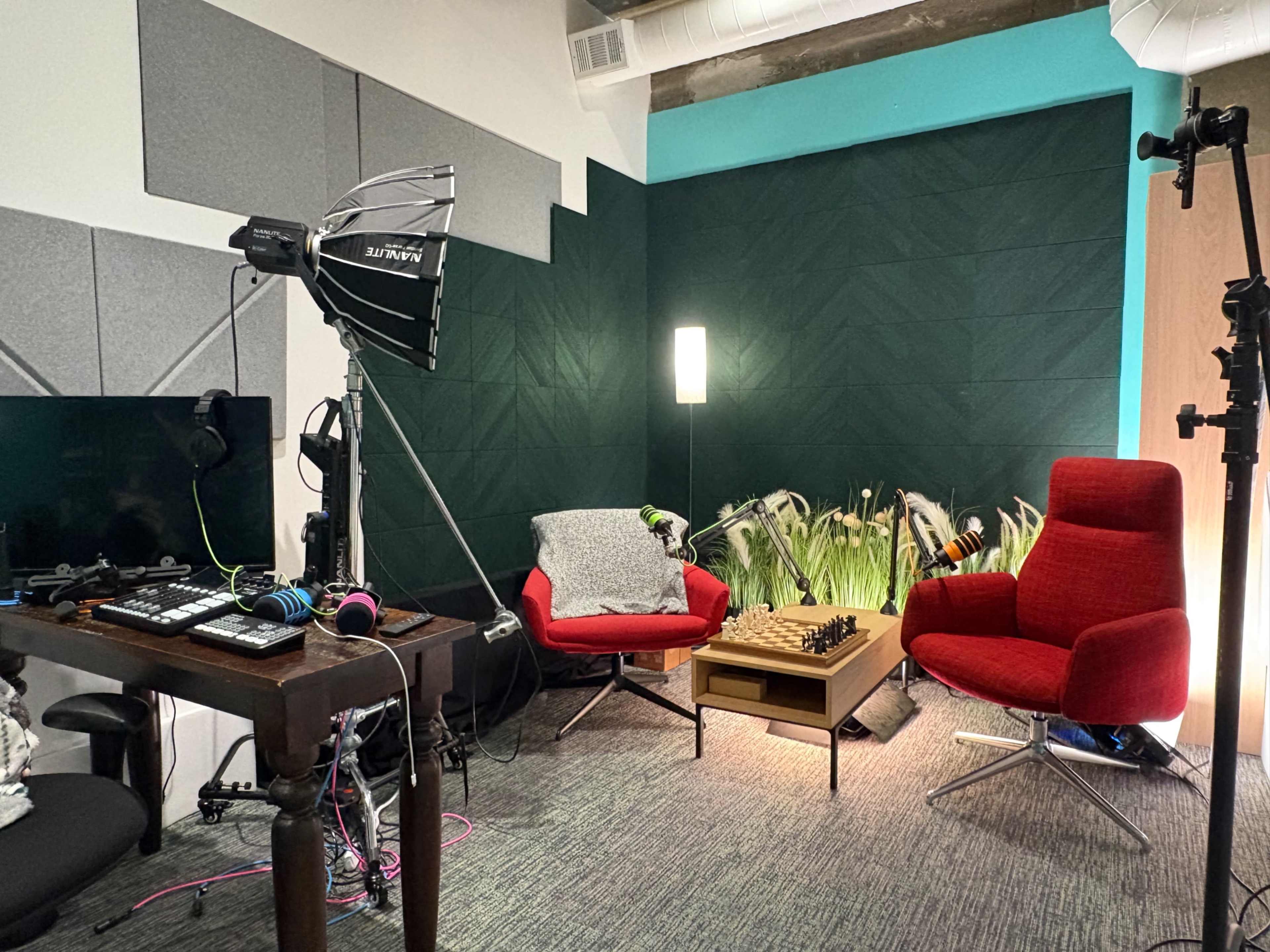 The image shows a small studio or workspace featuring two red chairs, a chessboard on a table, a computer monitor, and various lighting equipment.