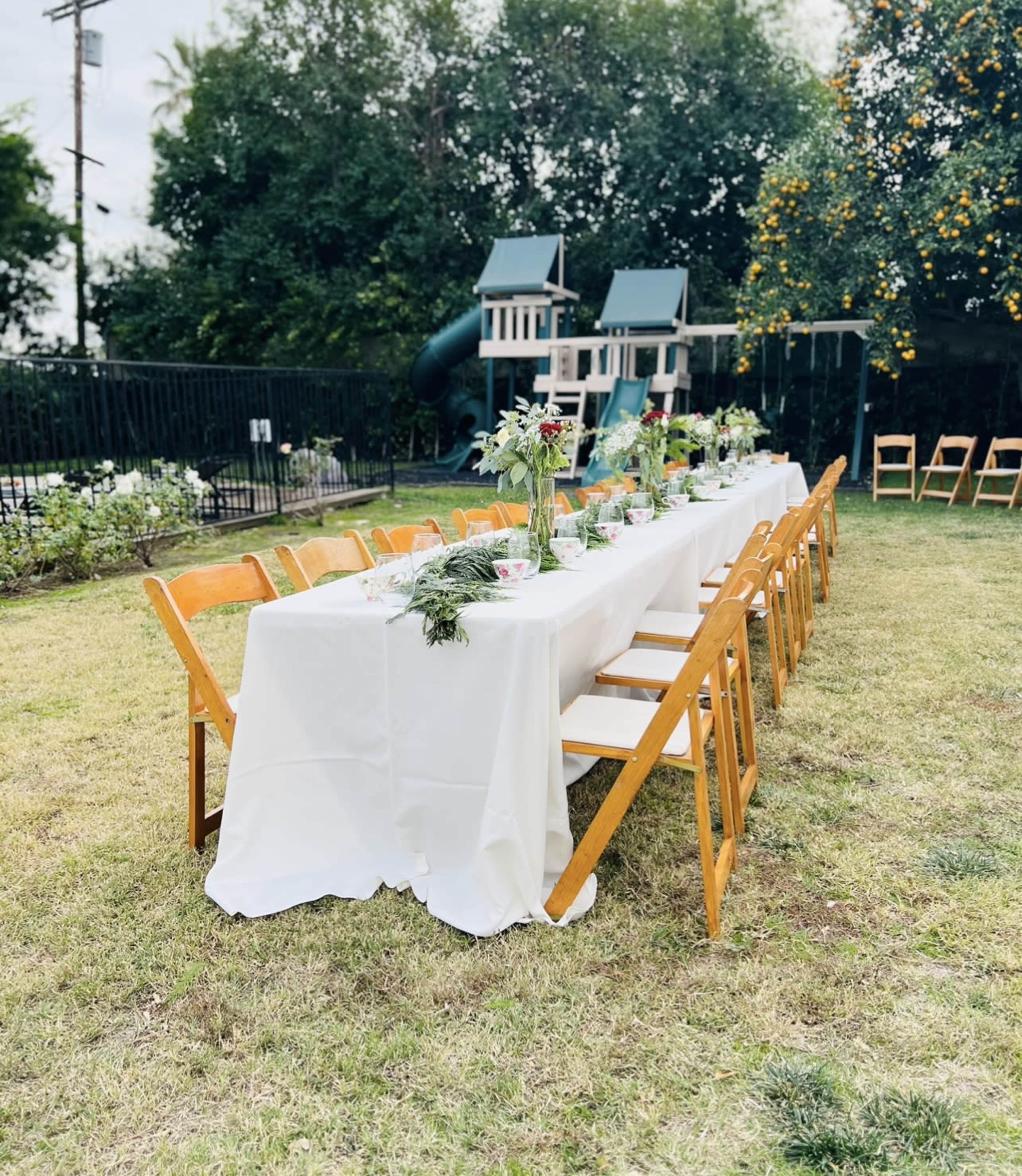 A long table covered in a white cloth is set up in a grassy backyard, surrounded by wooden chairs and facing a playground with a slide in the background.