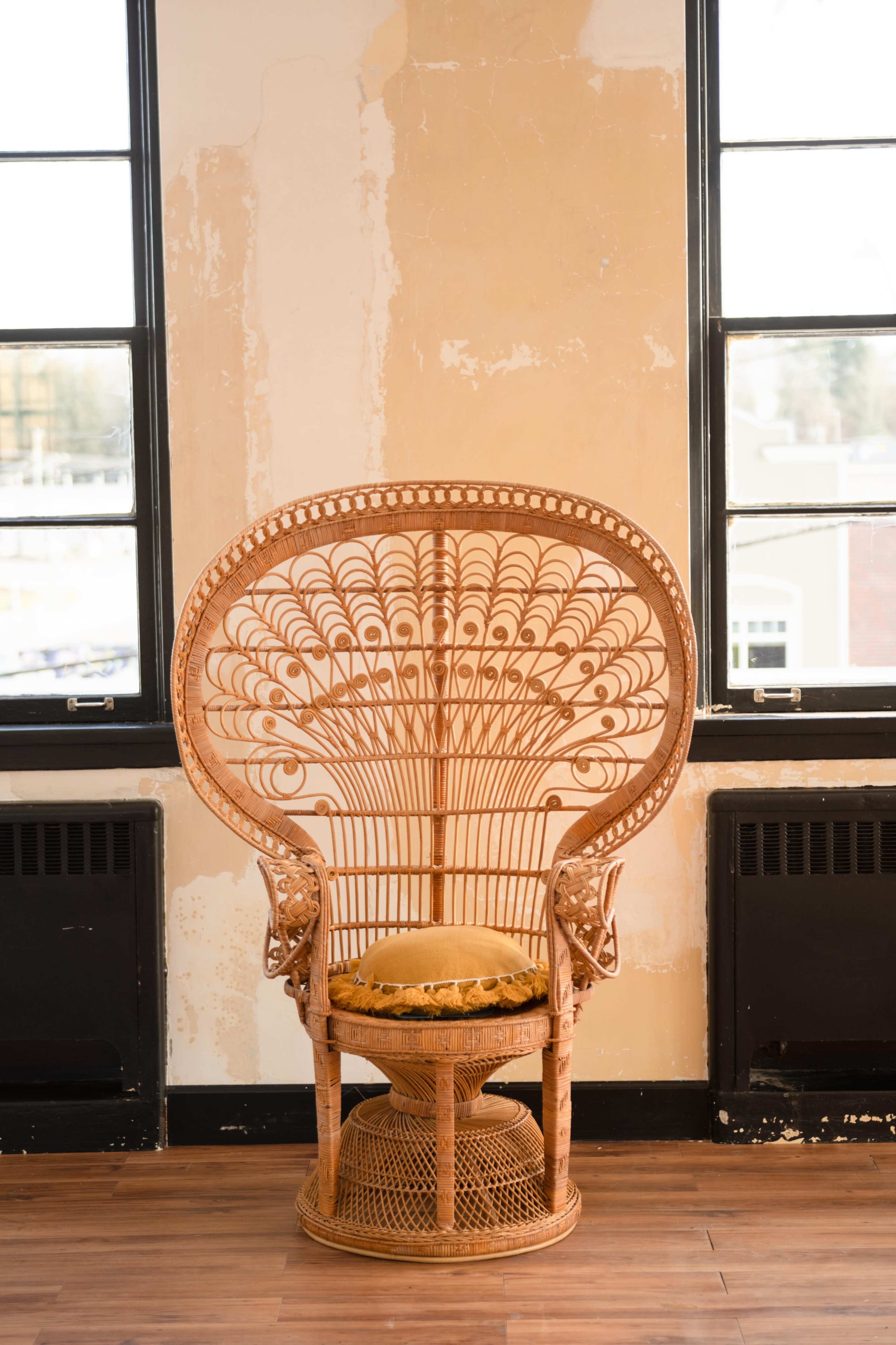A large, intricately designed rattan chair positioned in a room with exposed, peeling walls and large windows.