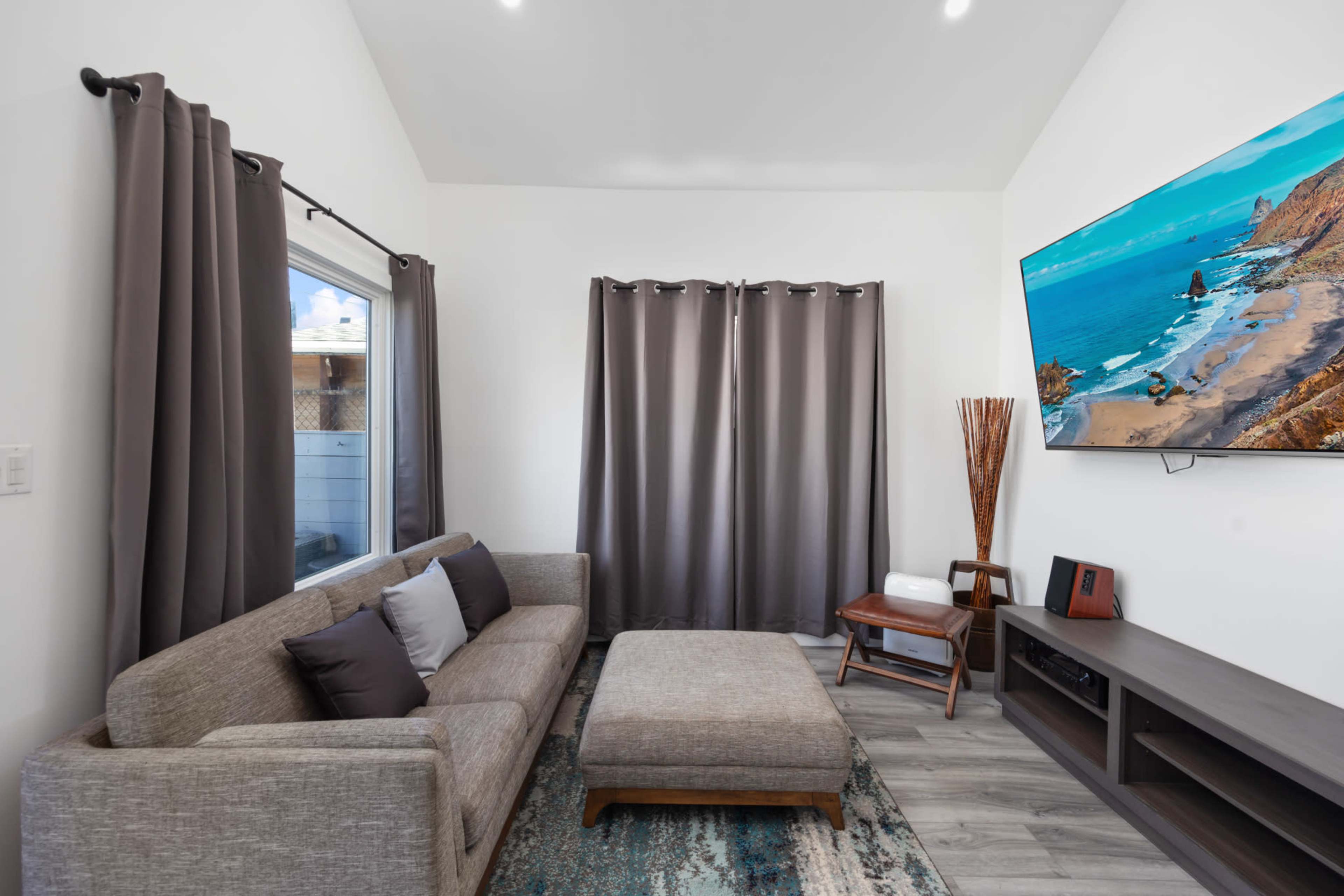 A modern living room features a gray sofa, an ottoman, curtains, and a wall-mounted television displaying a beach scene.