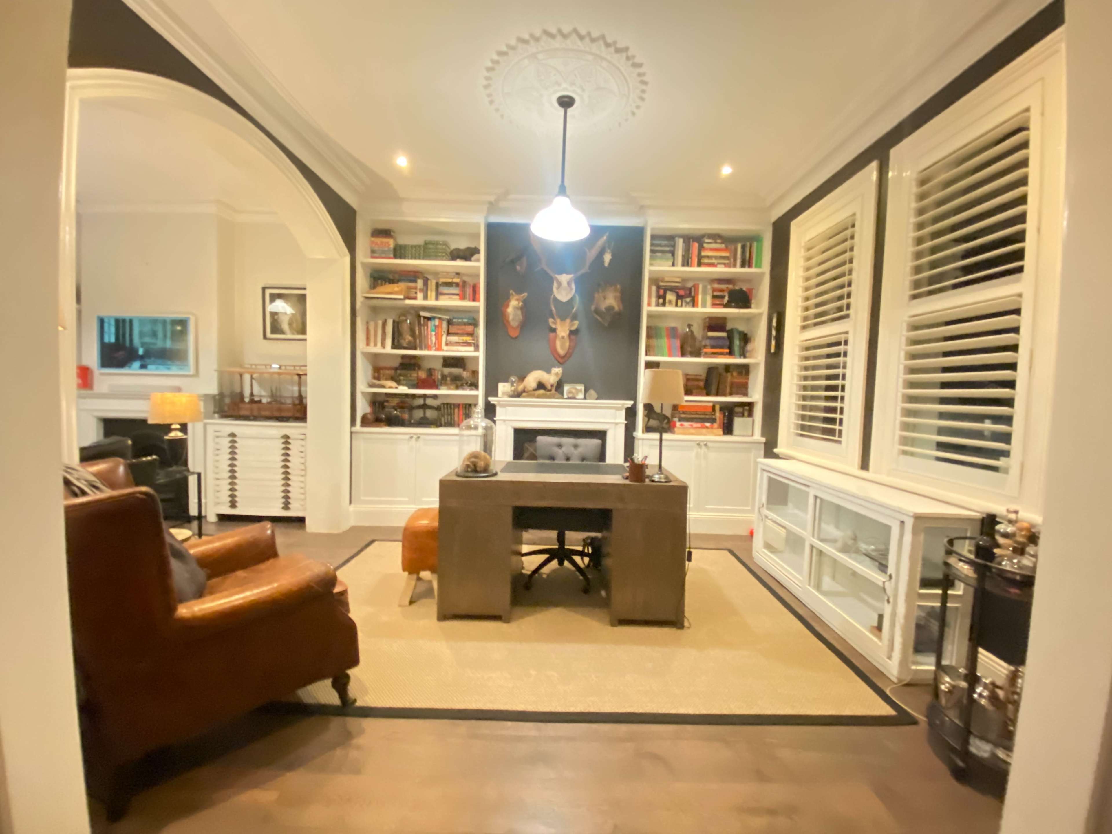 The image shows a stylish home office featuring a wooden desk, leather armchair, built-in bookshelves filled with books and decorative items, and a decorative wall with animal heads mounted.