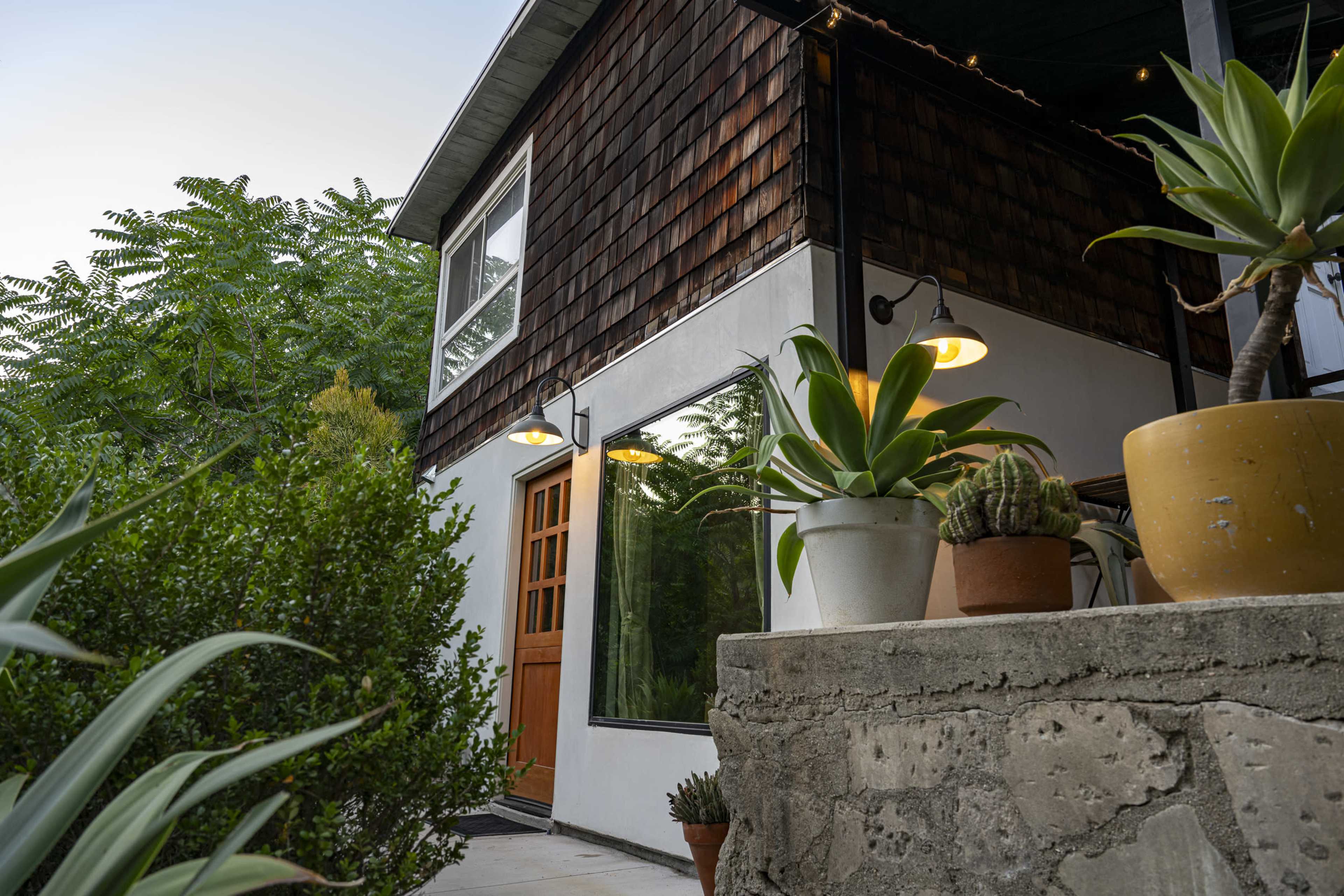 The image shows a modern house with wood shingles and a large window, surrounded by greenery and potted plants on a stone wall.