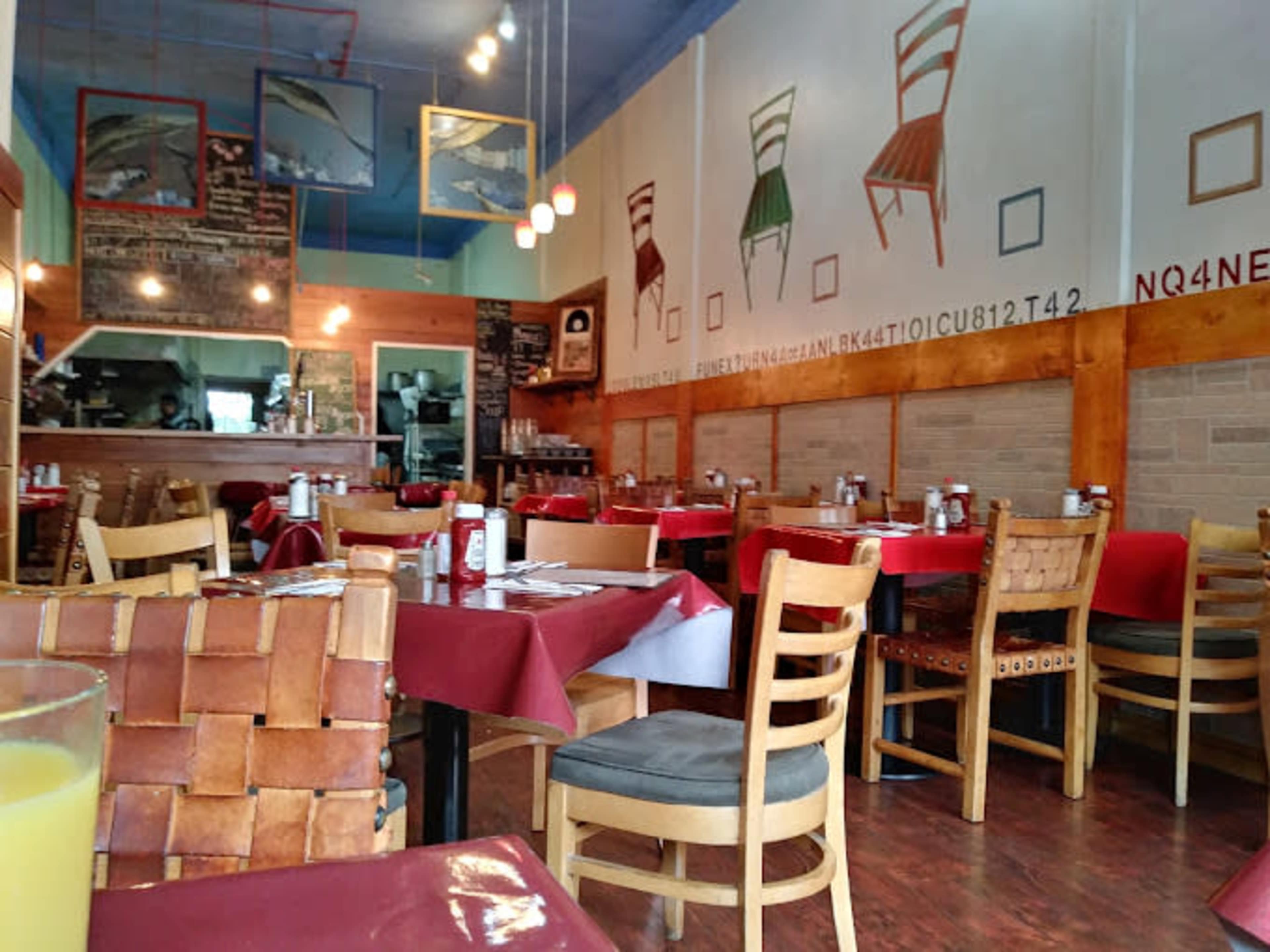The image shows a restaurant interior with tables set for dining, featuring wooden chairs, red tablecloths, and a wall decorated with framed items and a chalkboard menu.