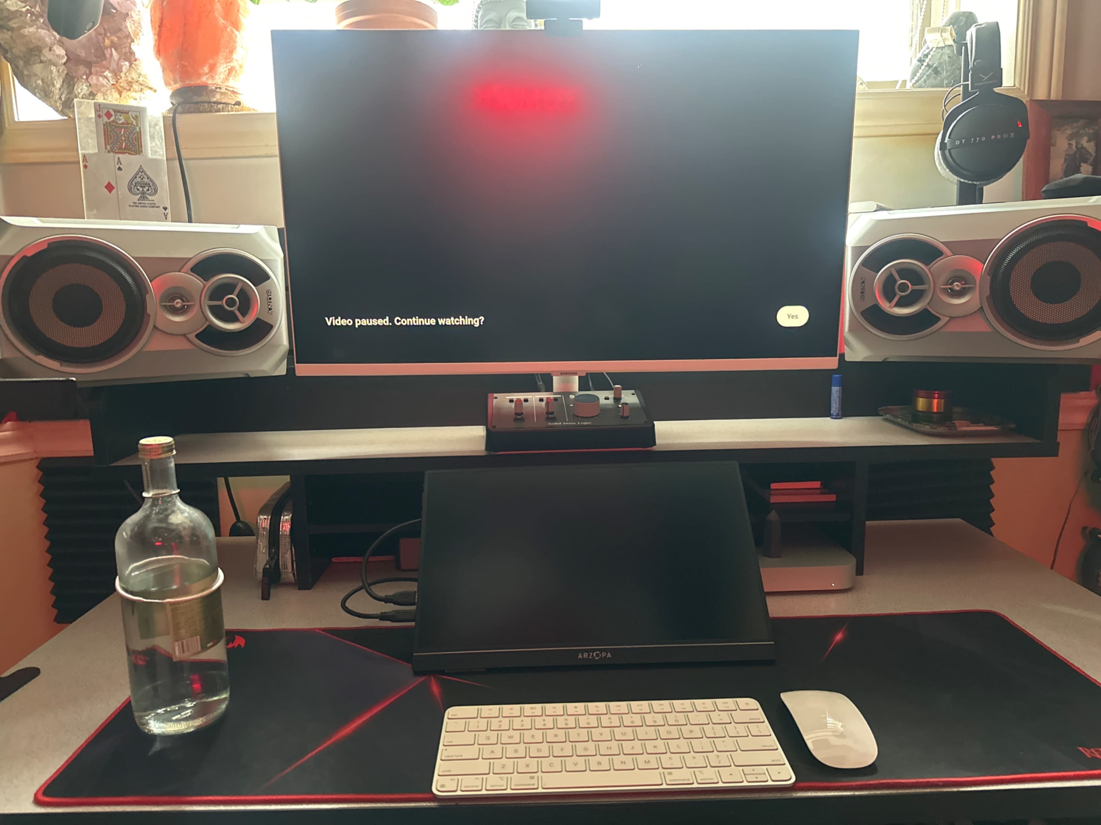 A clean desk setup features a large monitor displaying a paused video, flanked by two speakers, a small touchscreen device, a keyboard, and a bottle of water.