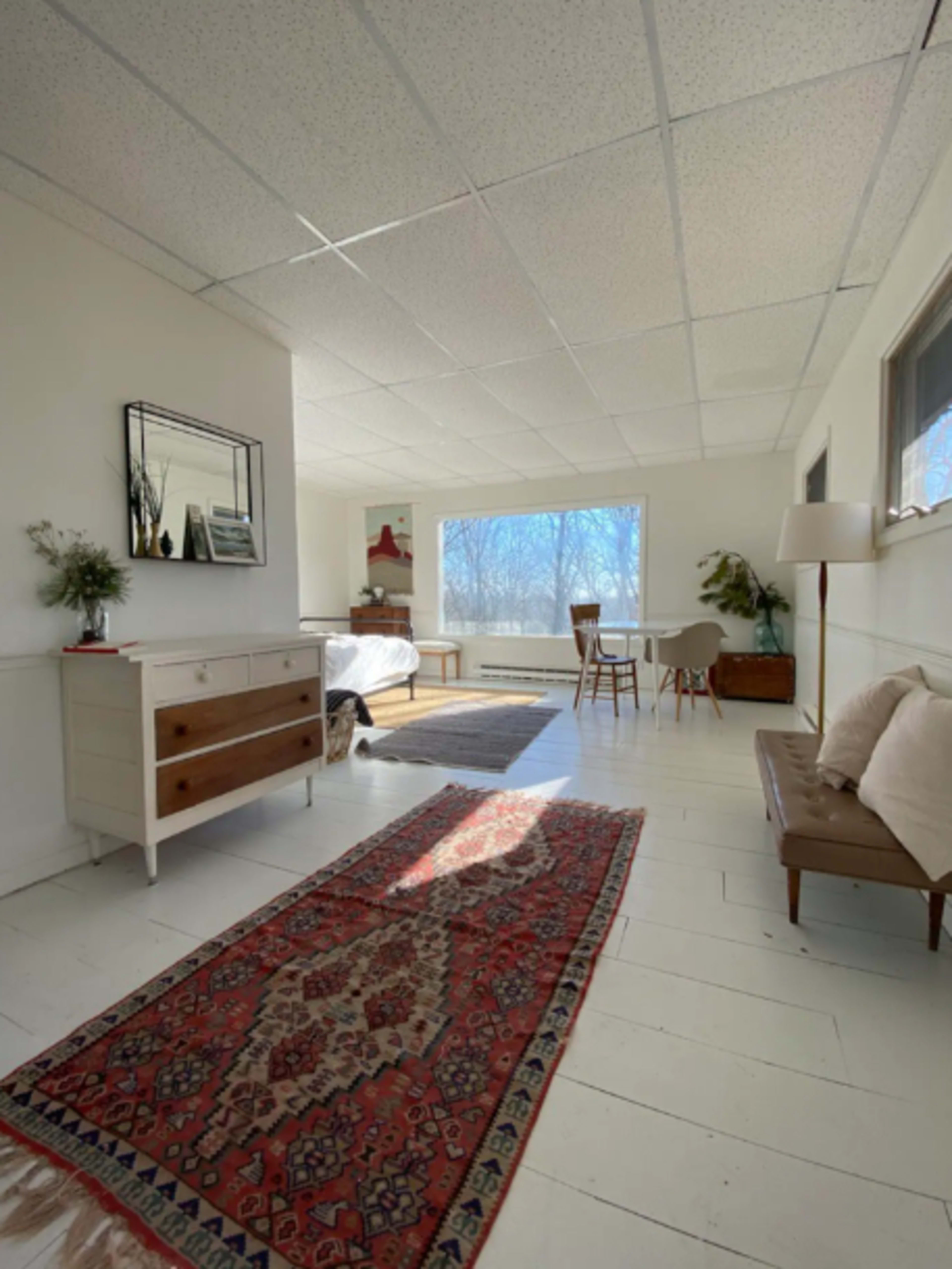 A bright, airy room with a wooden dresser, a bed in the background, and a rug on the white wooden floor, illuminated by natural light from large windows.