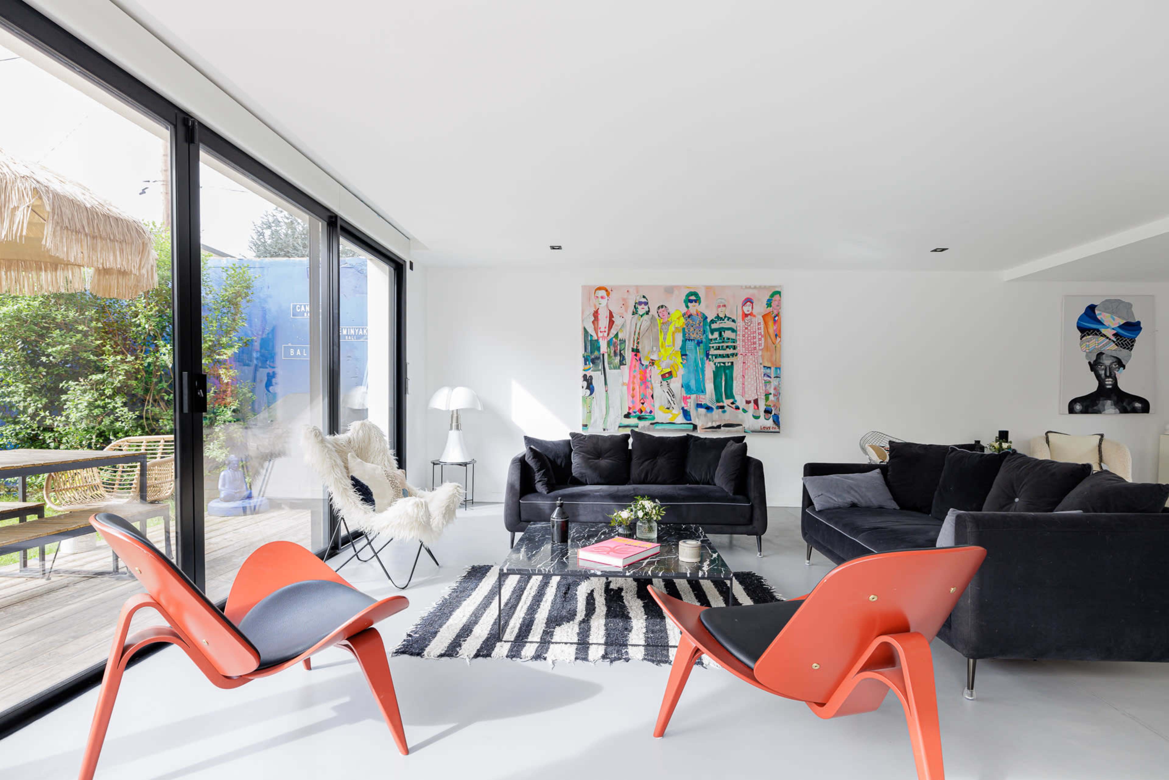 A modern living room features black sofas, colorful artwork on the wall, and a striped rug, with large glass doors opening to an outdoor space.
