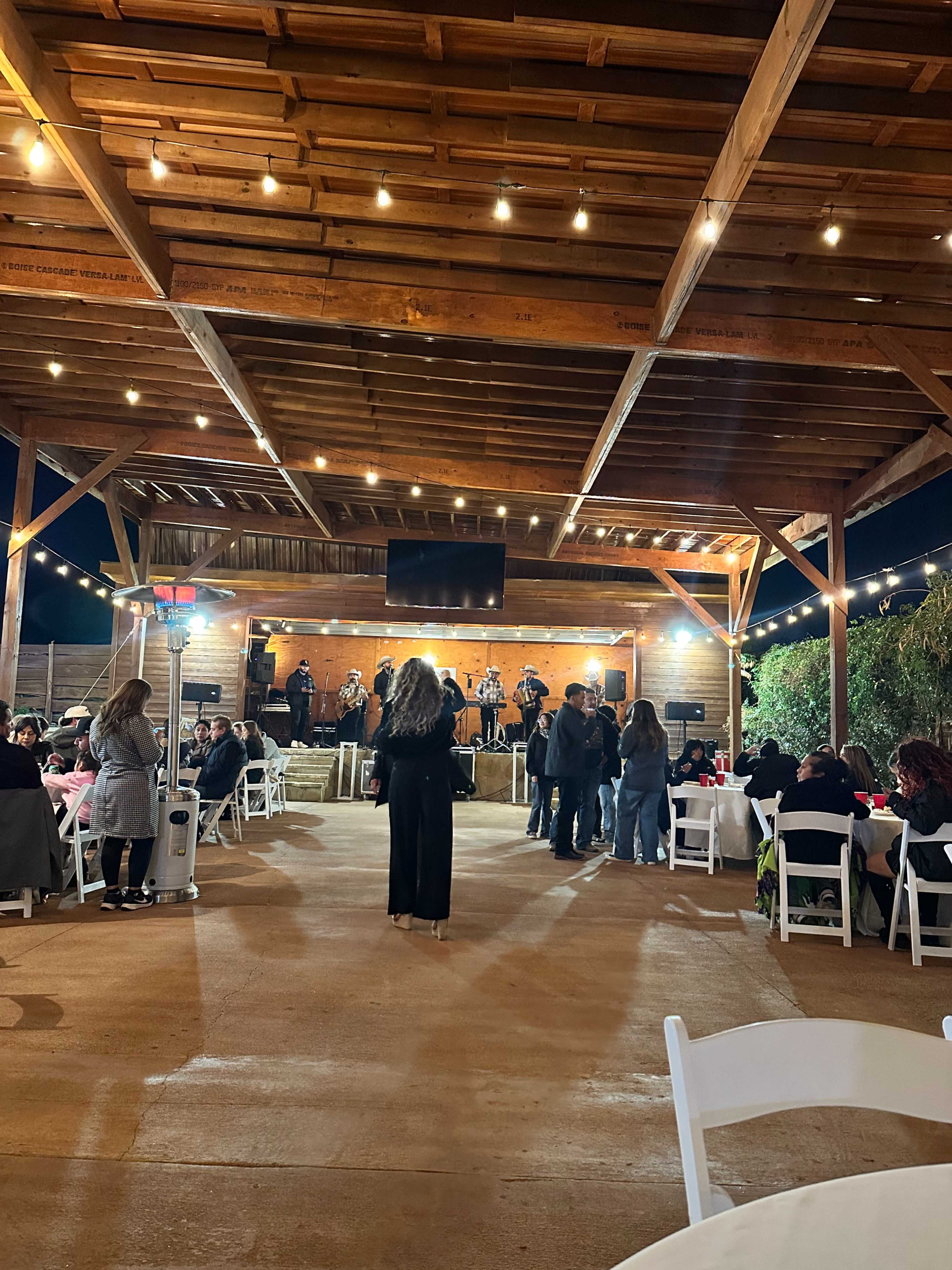 A band performs on a wooden stage while people gather at tables in a spacious, covered outdoor area illuminated by string lights.